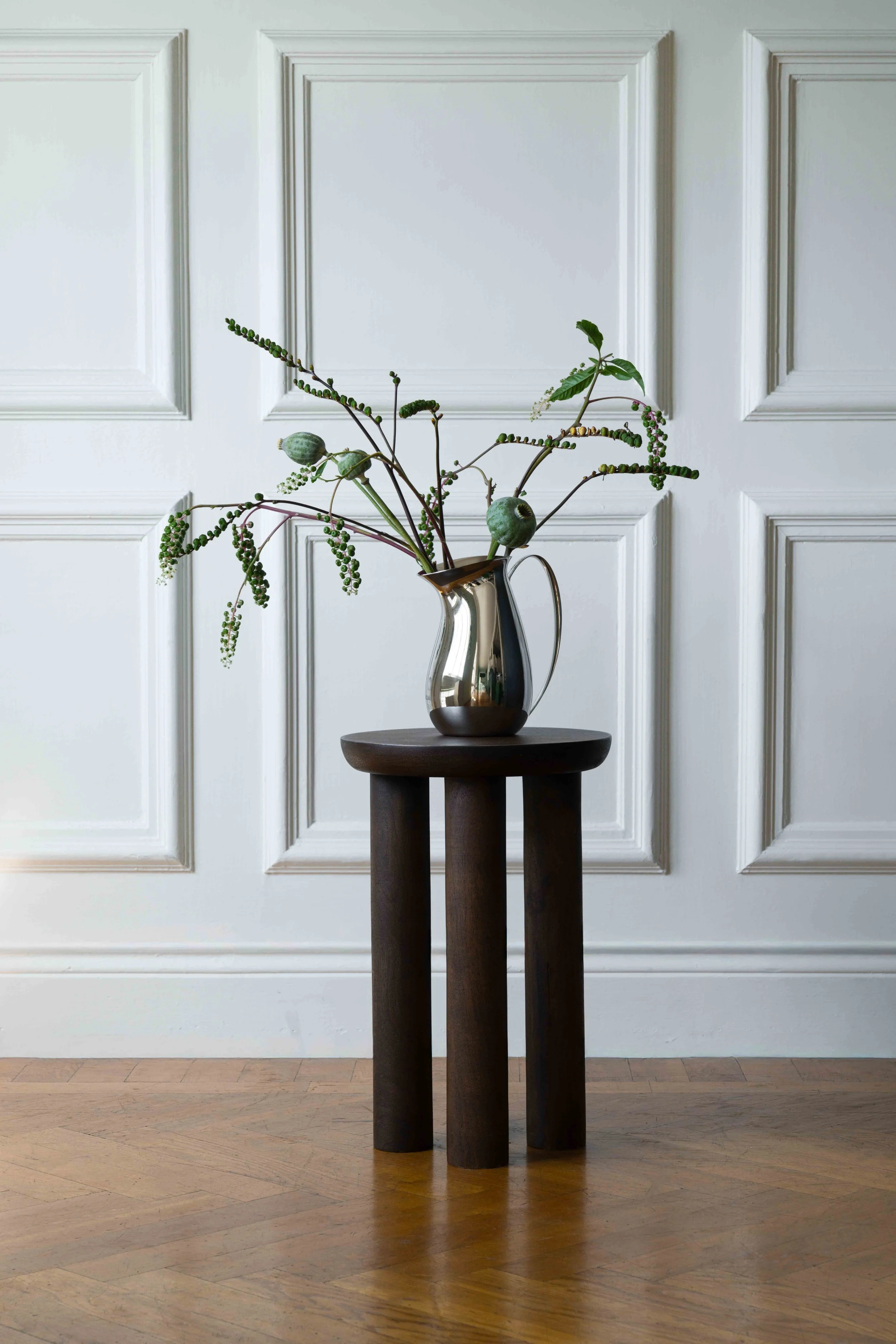 A modern still life with a metallic vase holding green plants and seed pods, placed on a dark wooden stand in front of white paneled wall and hardwood floor. Production by EKPR for Hilke Collection X Moomin.