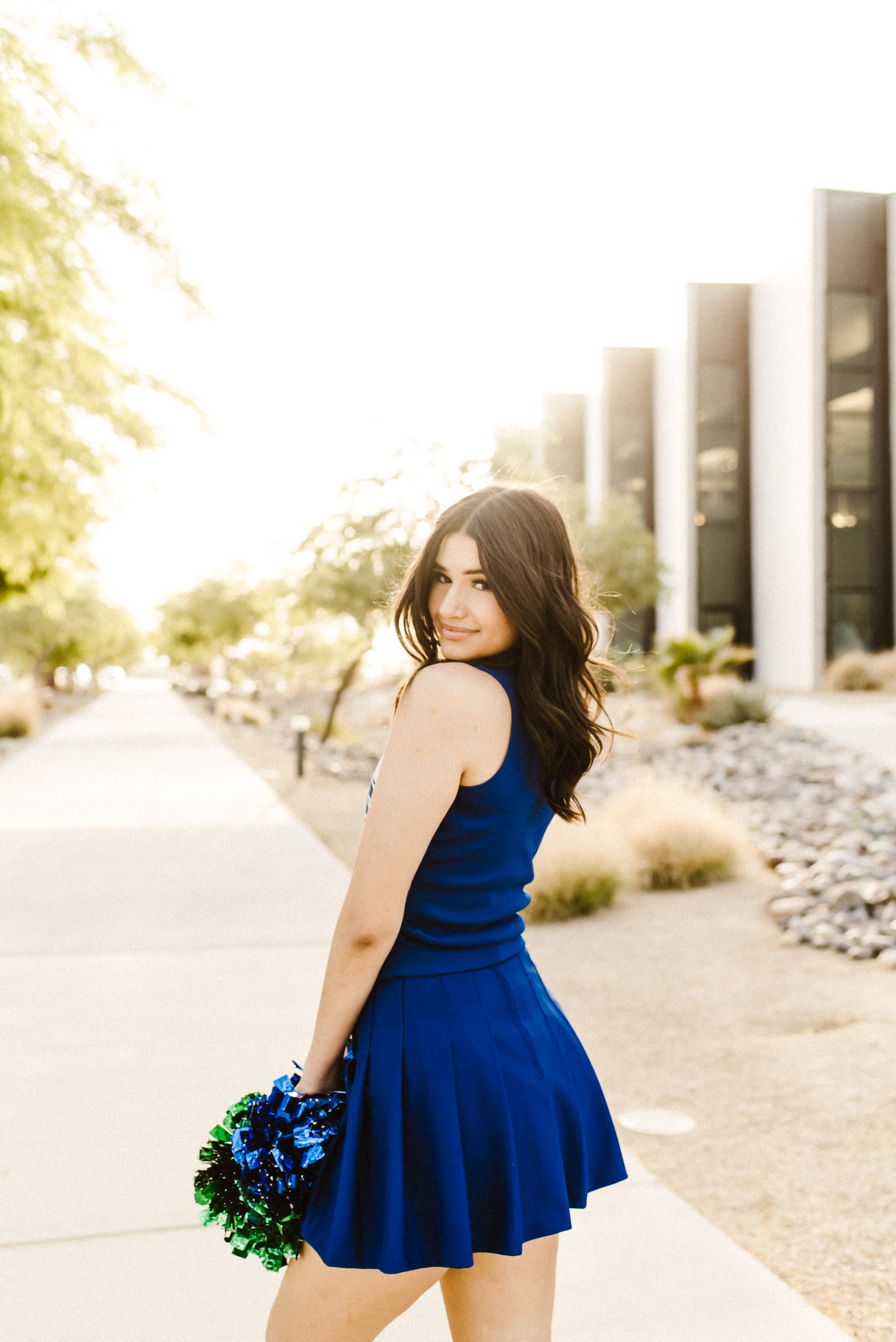 A young woman in a blue dress holding blue, green, and black pom-poms, standing outdoors in front of modern buildings and trees with sunlight shining behind her. El Paso Grad Photographer. El Paso Senior Photographer. El Paso Photographer.
