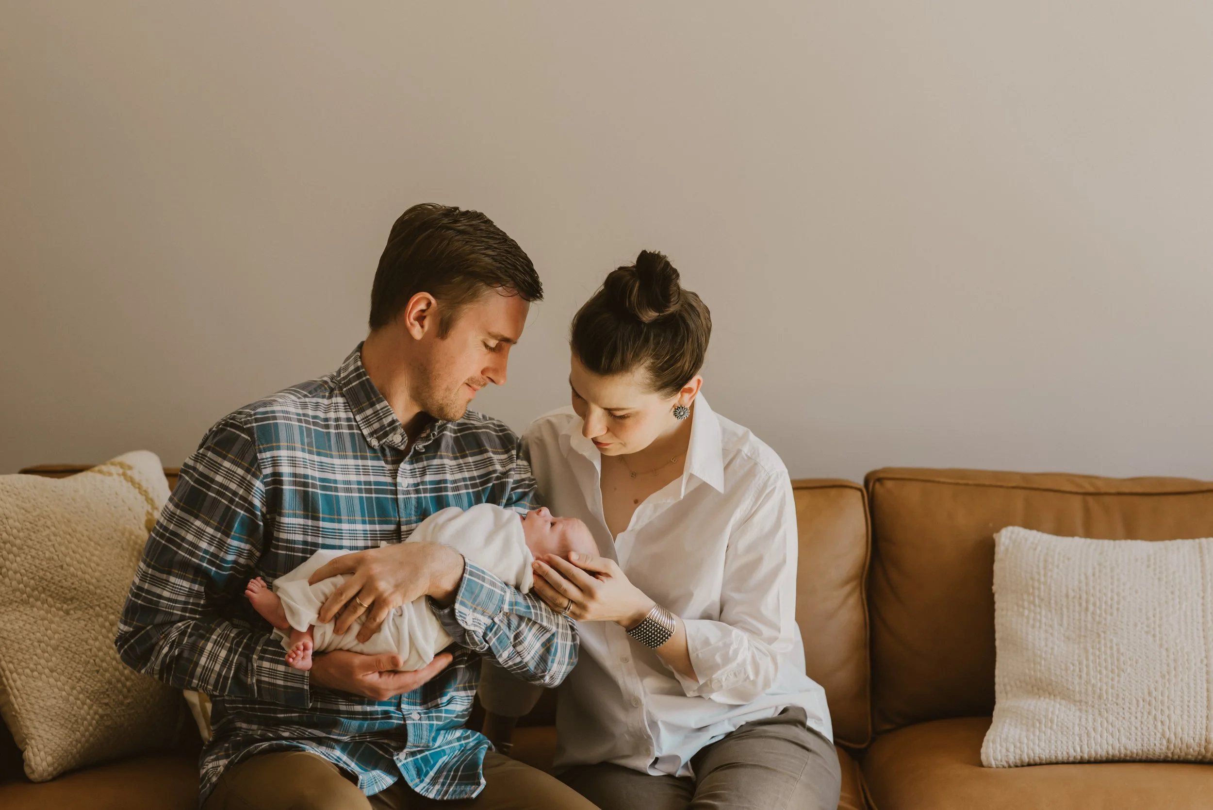 El Paso couple holding a newborn baby together in a living room with beige walls, sitting on a brown couch with beige cushions.  El Paso Newborn Photography. El Paso Lifestyle Photographer. El Paso Photographer.