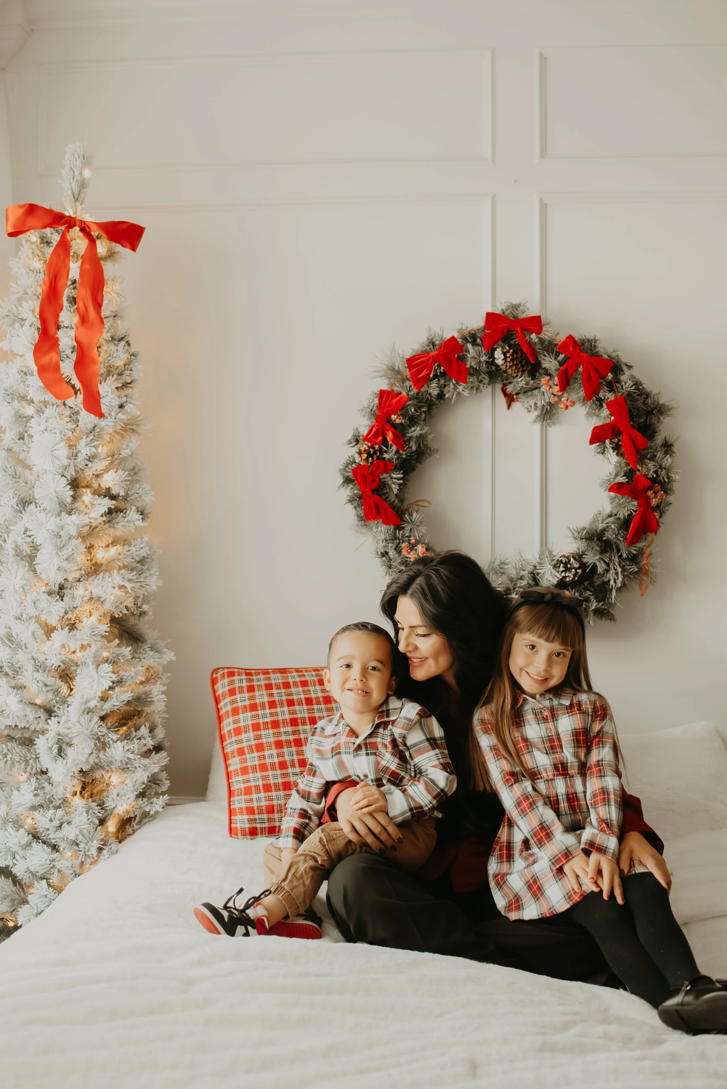 A woman with two children sitting on a bed in front of Christmas decorations, including a white Christmas tree adorned with a red ribbon and a wreath decorated with red bows, pinecones, and berries.