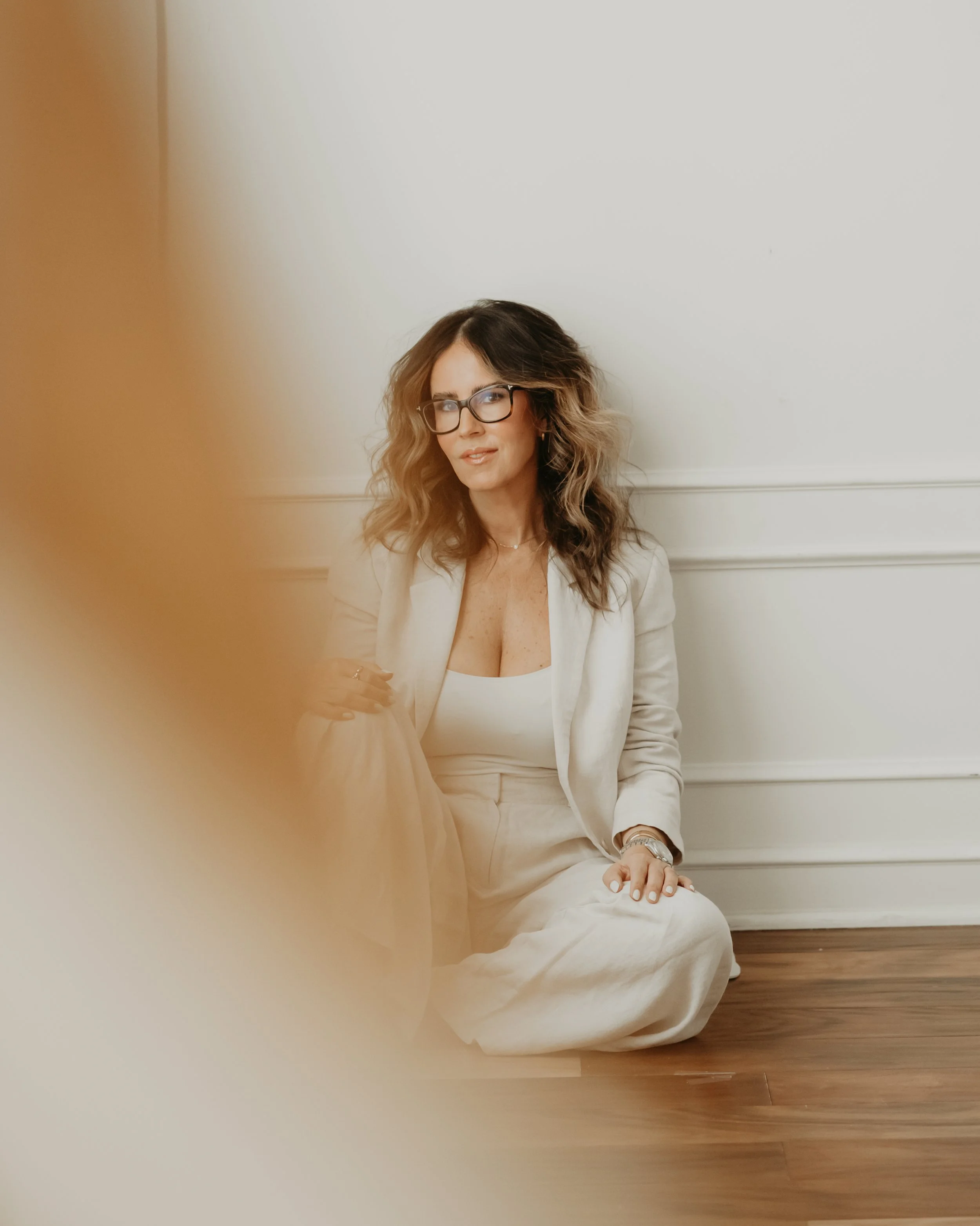 Woman with wavy brown hair, wearing glasses, a light beige blazer, and matching pants, sitting on the floor against a white wall with wooden flooring.