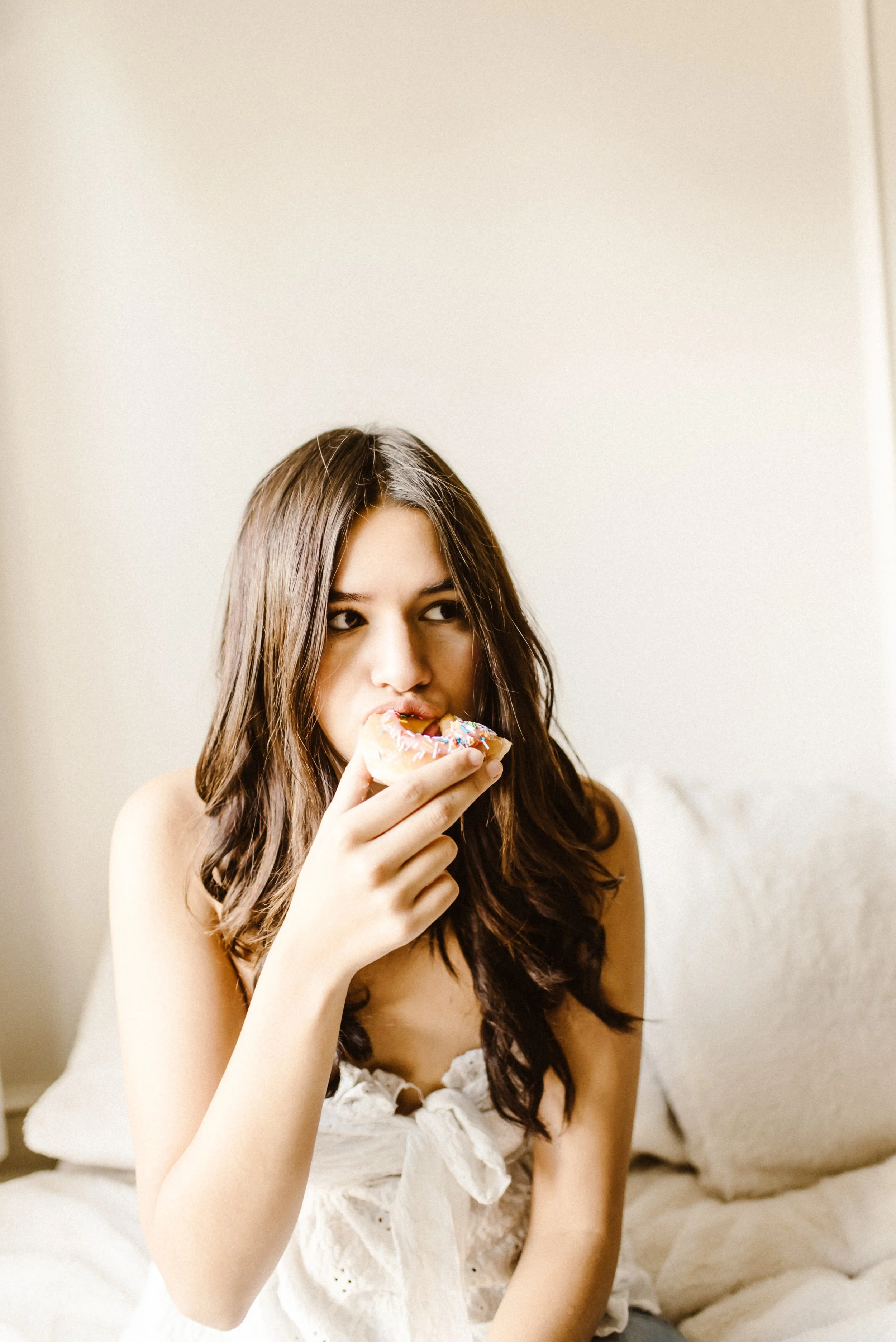 A young woman with long brown hair and light skin, wearing a white dress, is sitting on a bed, eating a donut with colorful sprinkles.
