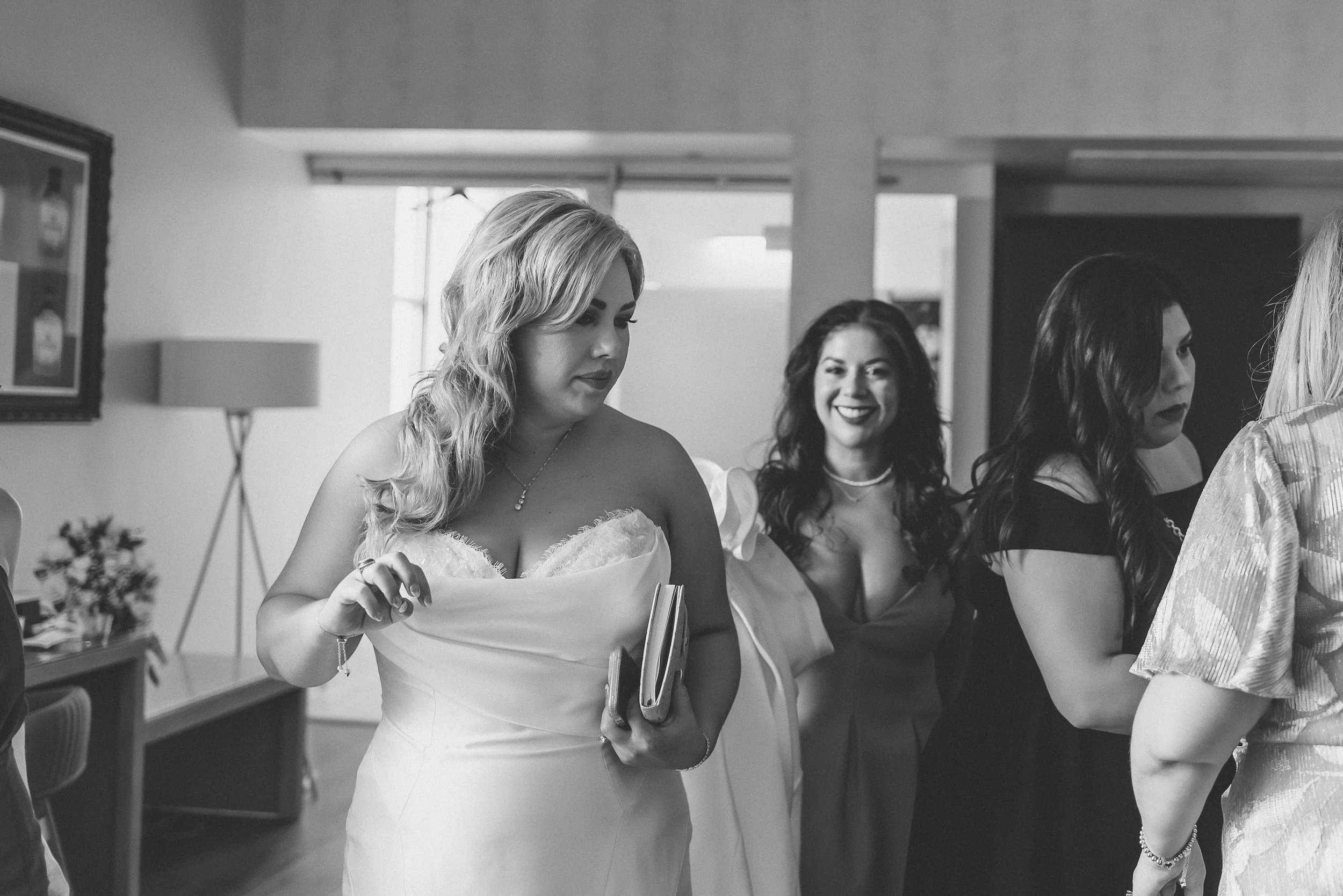 A group of women at a social gathering, with one woman in a white dress holding a book, smiling, while the others converse in a dimly lit room.