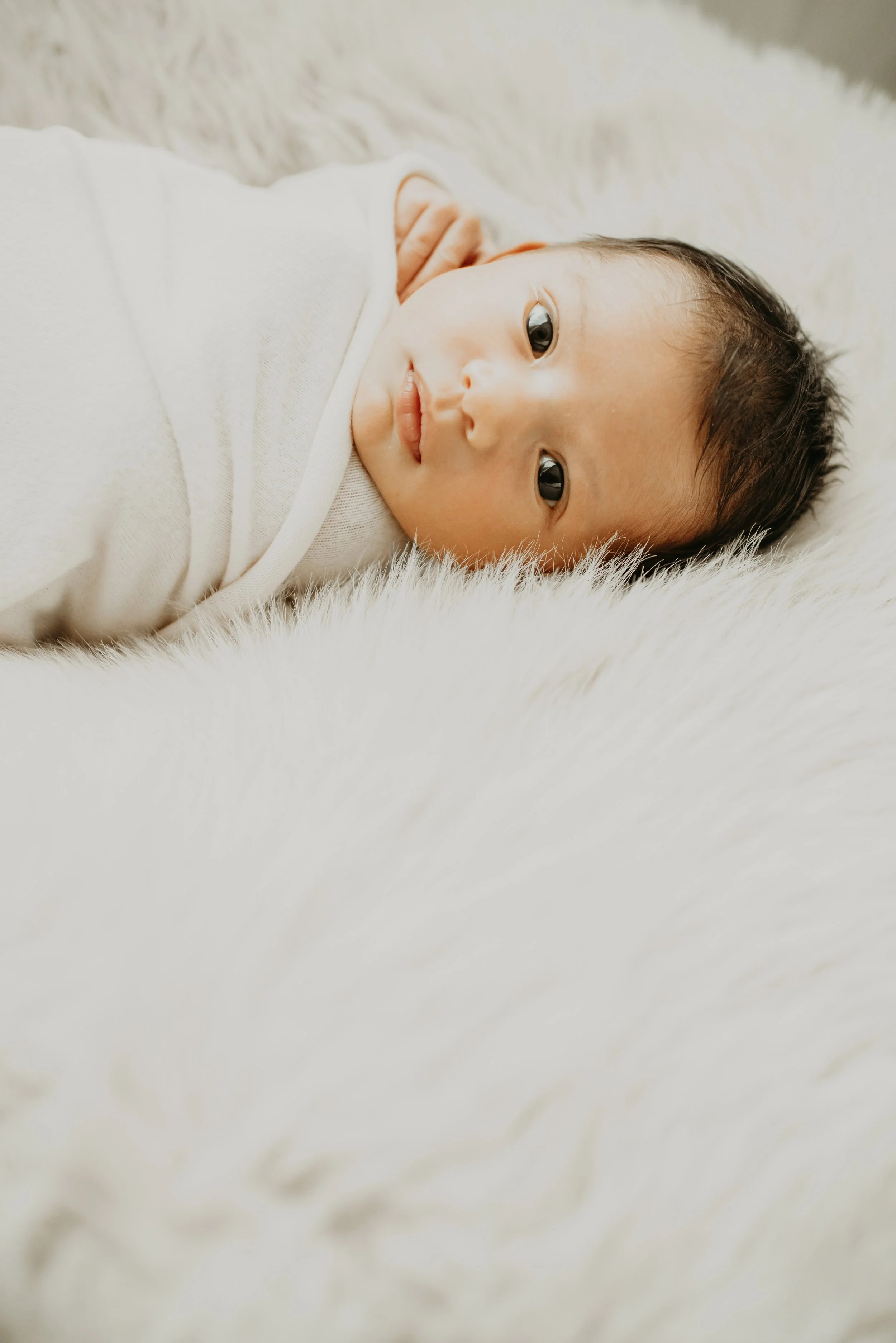 A baby laying on a soft, fluffy white surface, wearing a white onesie, looking at the camera with wide eyes.