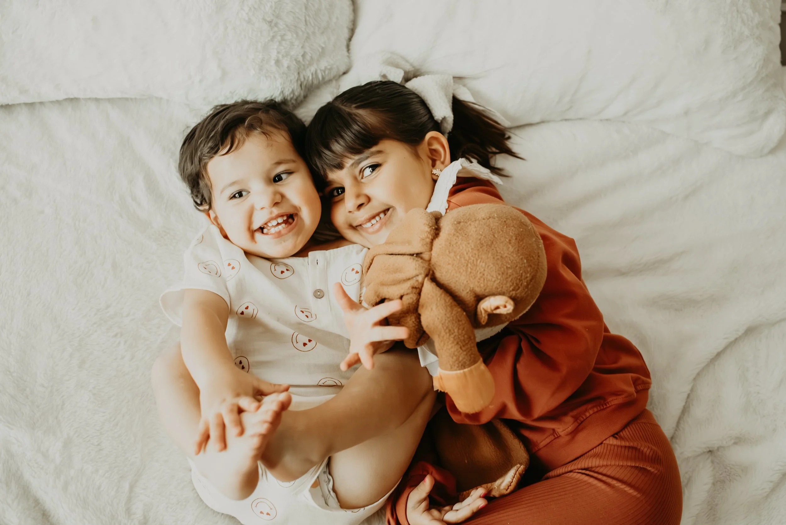 A young woman and a little boy lying on a white blanket, smiling and hugging each other, with the boy holding a brown teddy bear.