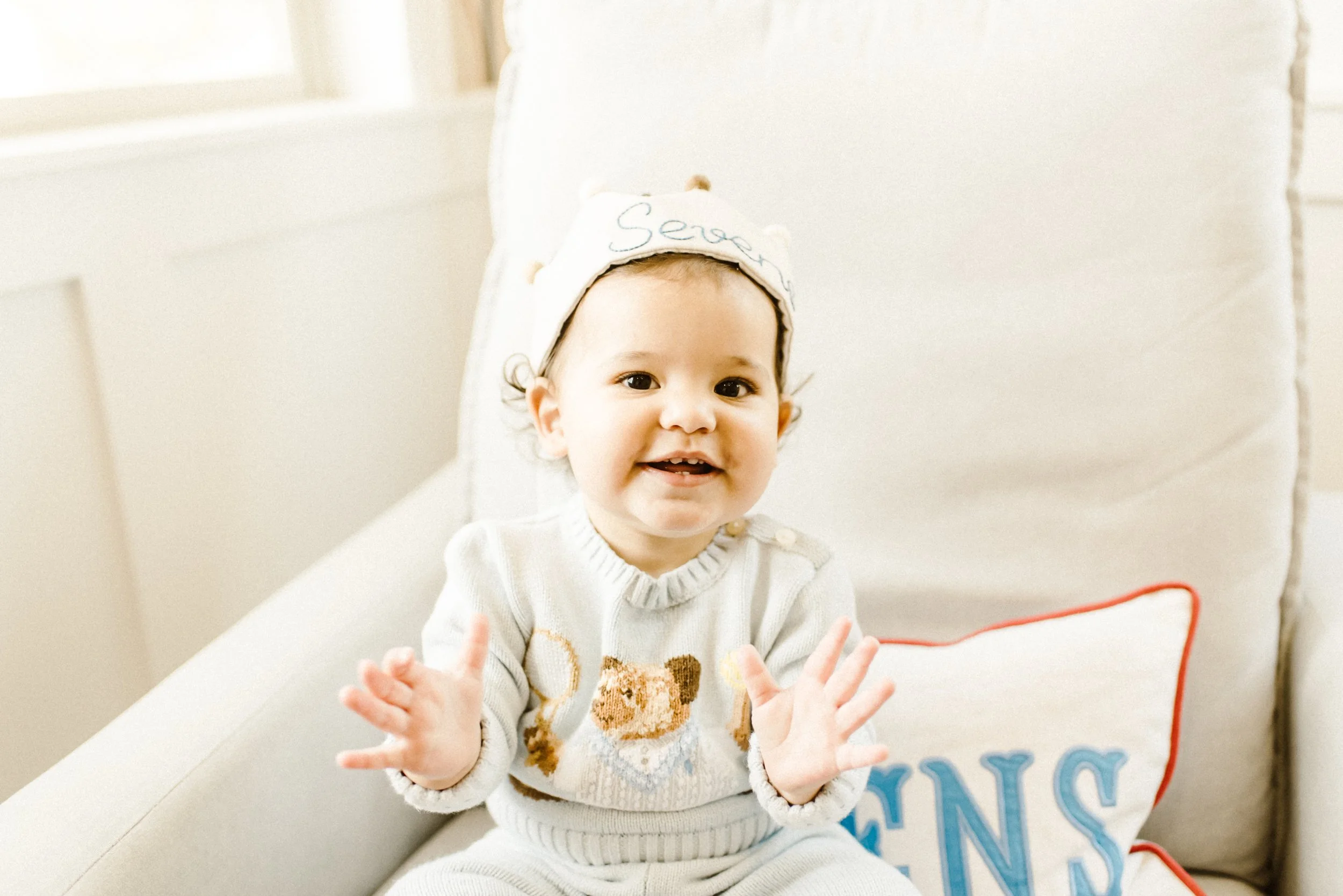 A smiling young child with light skin and dark hair, wearing a white sweater with a dog pattern and a white hat with embroidered writing, sitting on a white couch and reaching out with their hands.