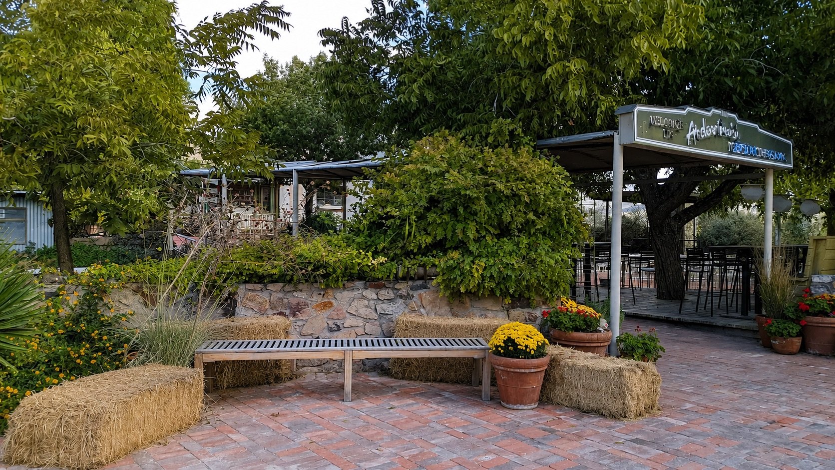 Outdoor patio area with straw bale benches, potted flowers, and lush green trees. There is a sign with partially visible text, and a covered dining space with tables and chairs in the background.