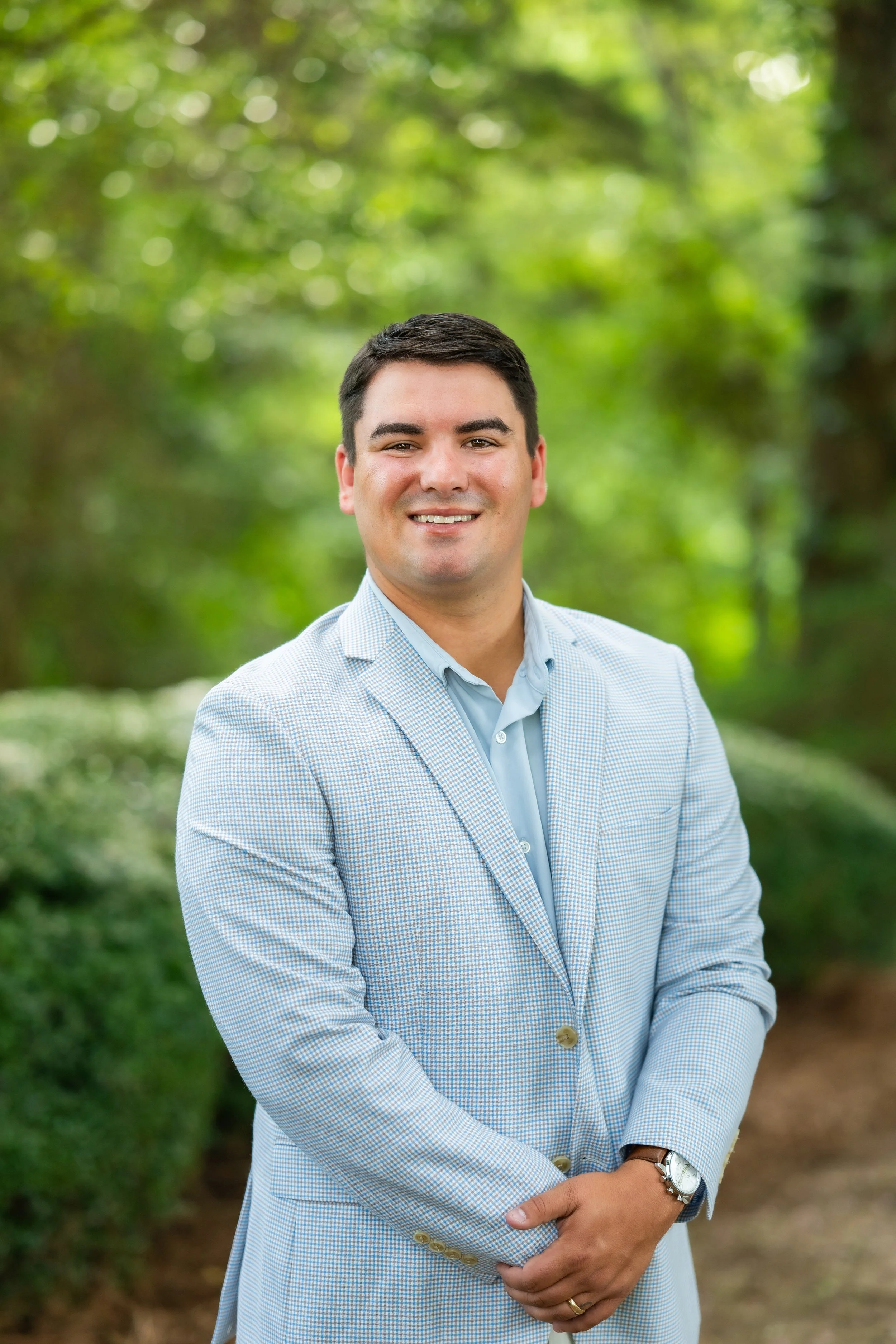 A young man in a light blue checkered blazer and light blue shirt, standing outdoors against a background of green trees and blurred foliage, smiling and looking at the camera with arms crossed in front of him.