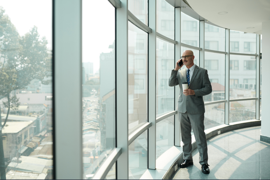 Businessman in gray suit holding a coffee cup and talking on a mobile phone near large curved glass windows in a modern office building.