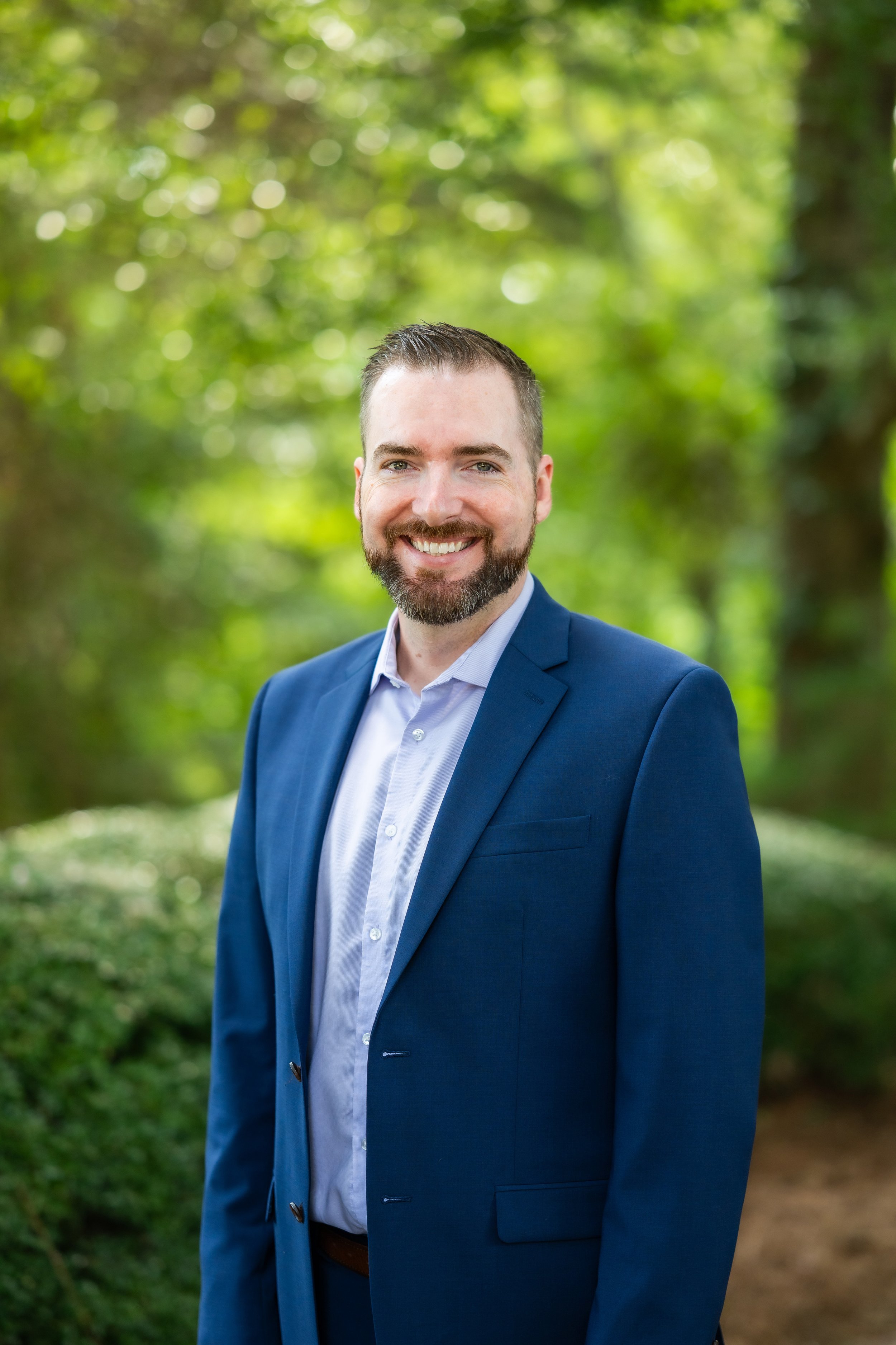 A man with a beard and short hair, wearing a blue suit jacket and a light blue collared shirt, smiling outdoors with a blurred green forest background.