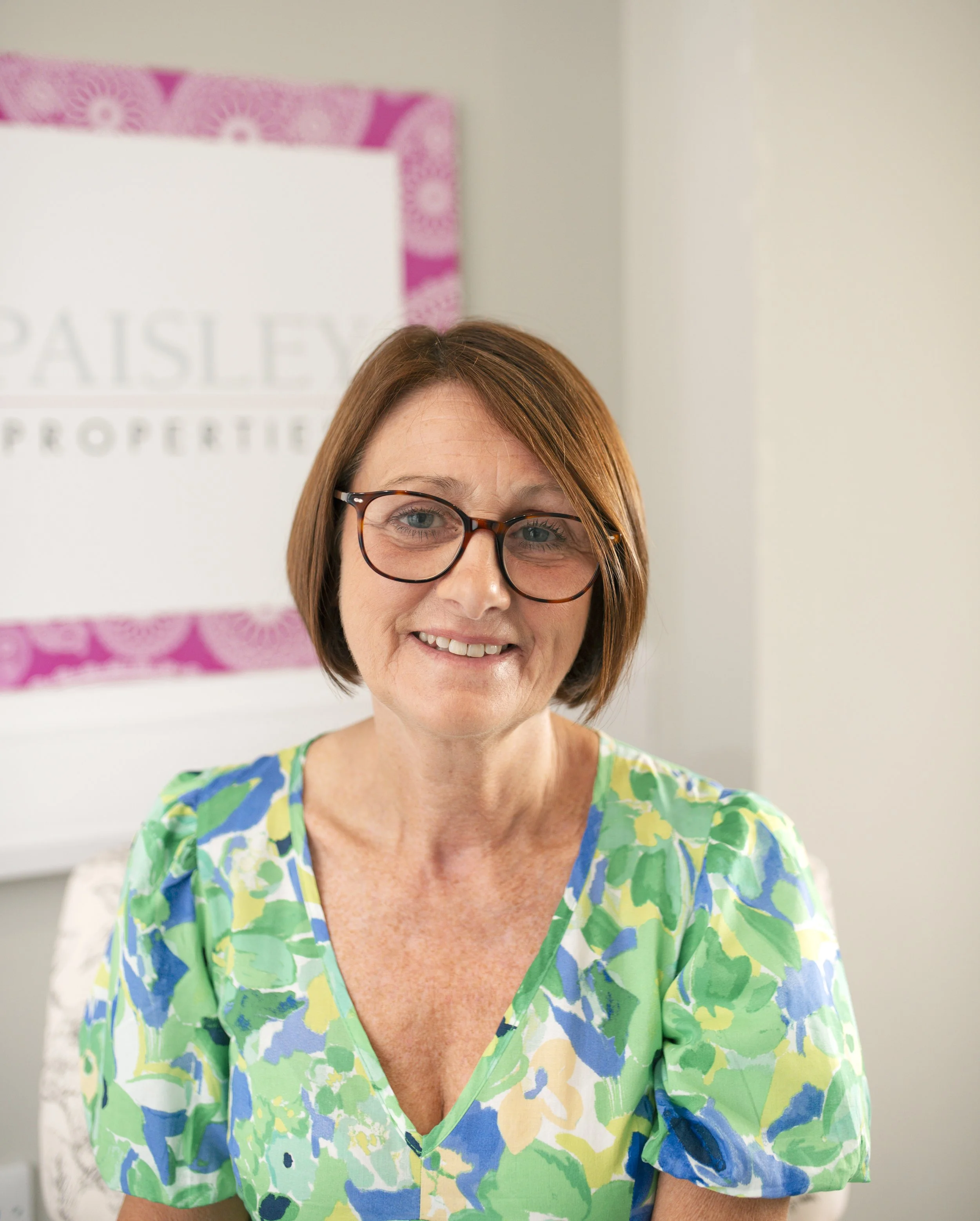 A woman with red hair and glasses smiling in an office, wearing a colorful green, blue, and yellow floral top.