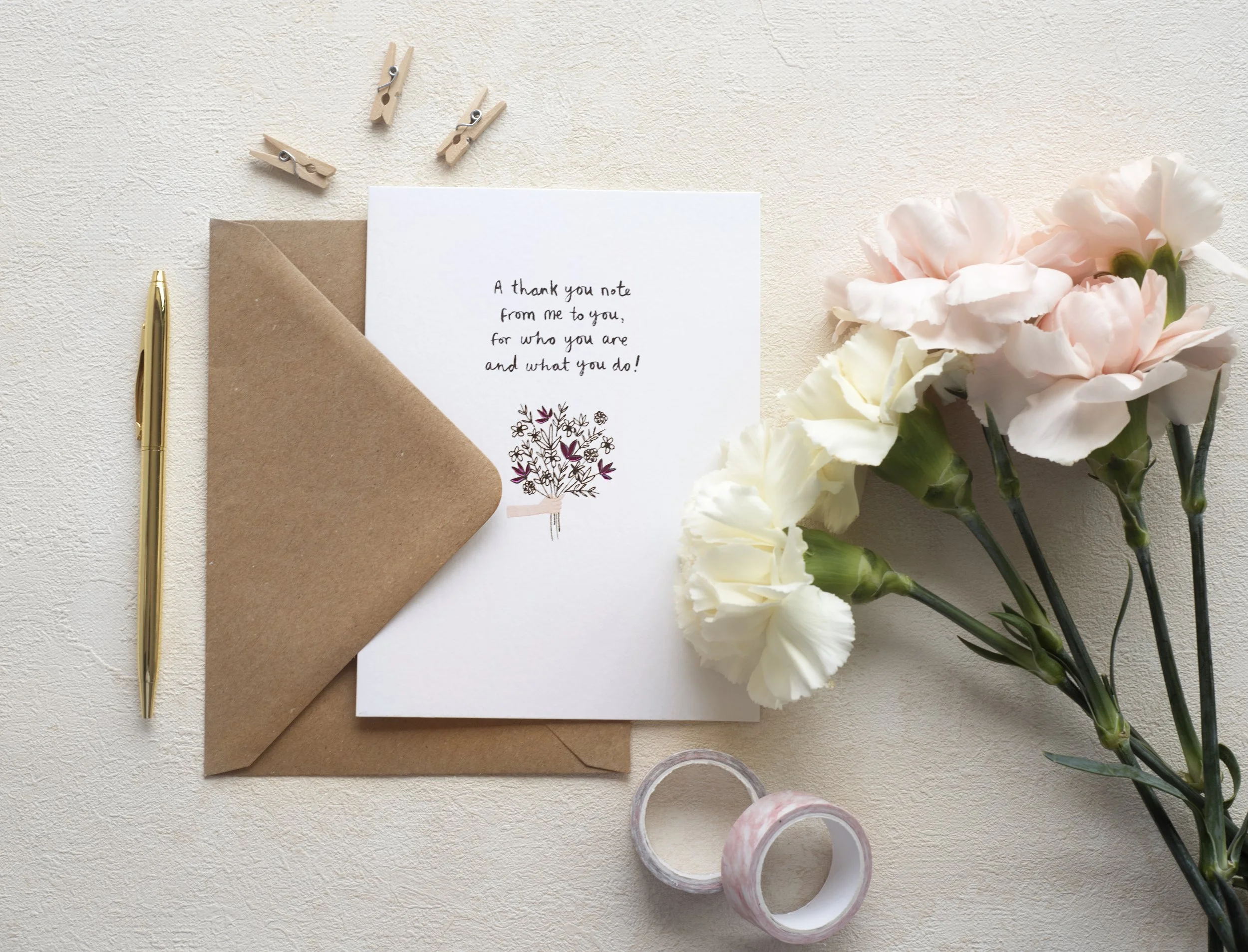 A thank you note card with a bouquet of pale pink and white flowers, a brown envelope, a gold pen, small clothespins, and decorative washi tape on a light textured surface.