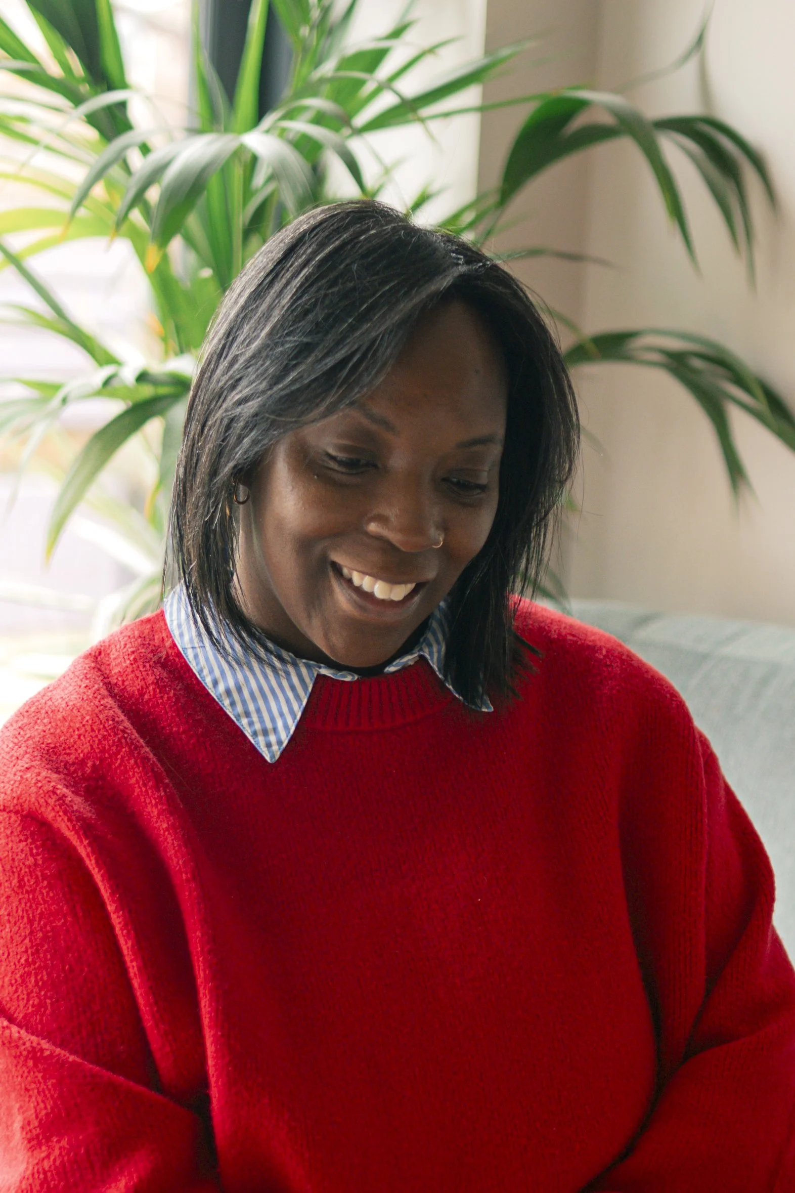 A woman sits on a couch wearing a red sweater over a blue striped shirt, smiling while looking down, with green houseplants in the background.