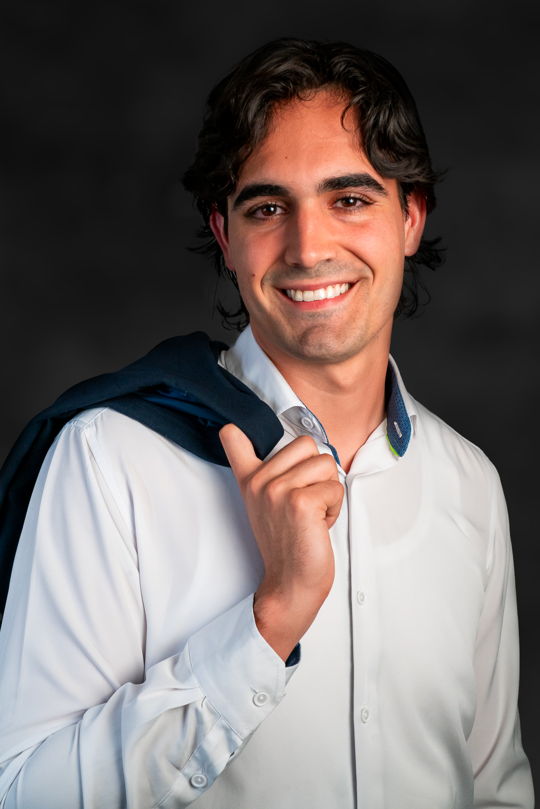 Young man with dark hair and a bright smile, holding a dark jacket over his shoulder, wearing a white button-up shirt, against a dark background.