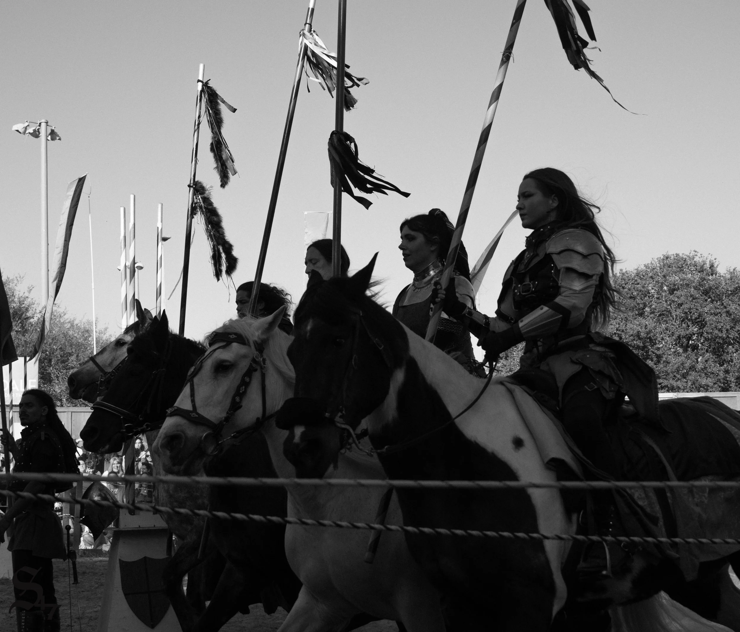 People dressed in medieval armor riding horses, holding flags or banners, at an outdoor event.