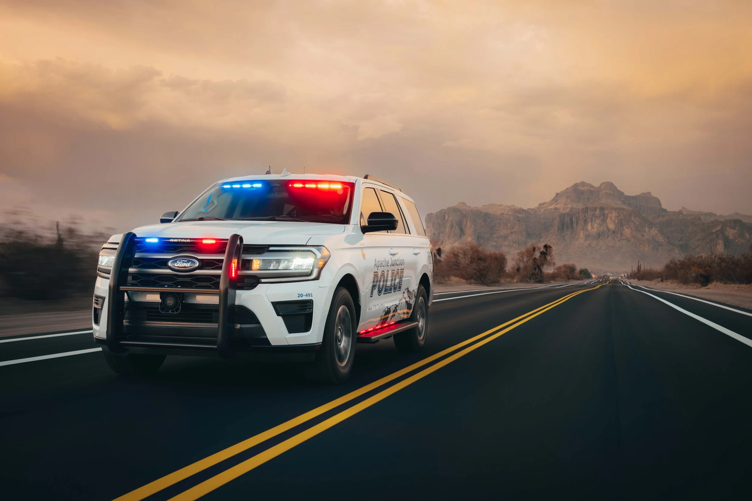 Two police SUVs with flashing red and blue lights driving on a rural road during dusk.