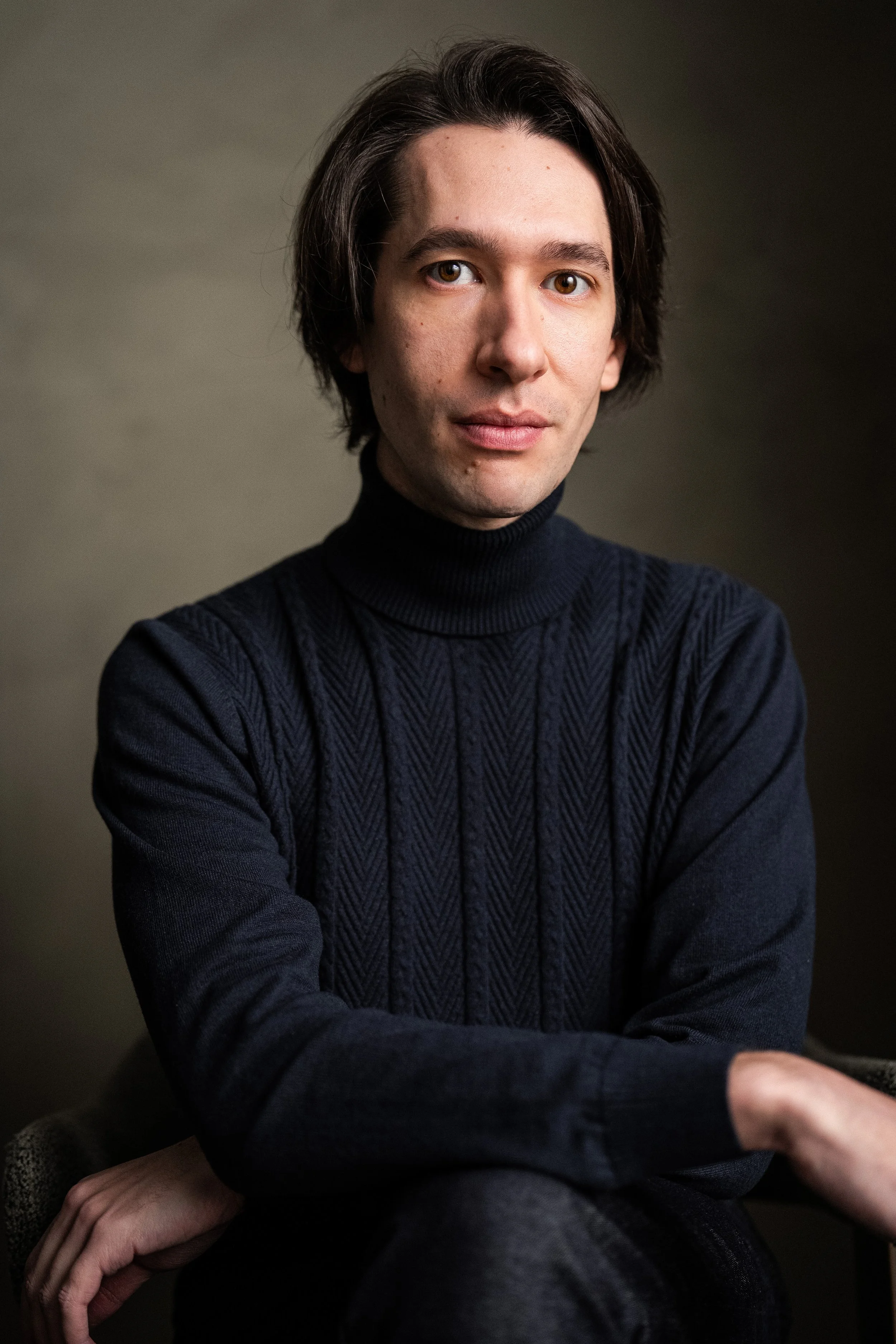Theatre actor Christian Pascolutti wearing a dark blue turtleneck sweater, sitting with arms crossed against a plain, neutral background.