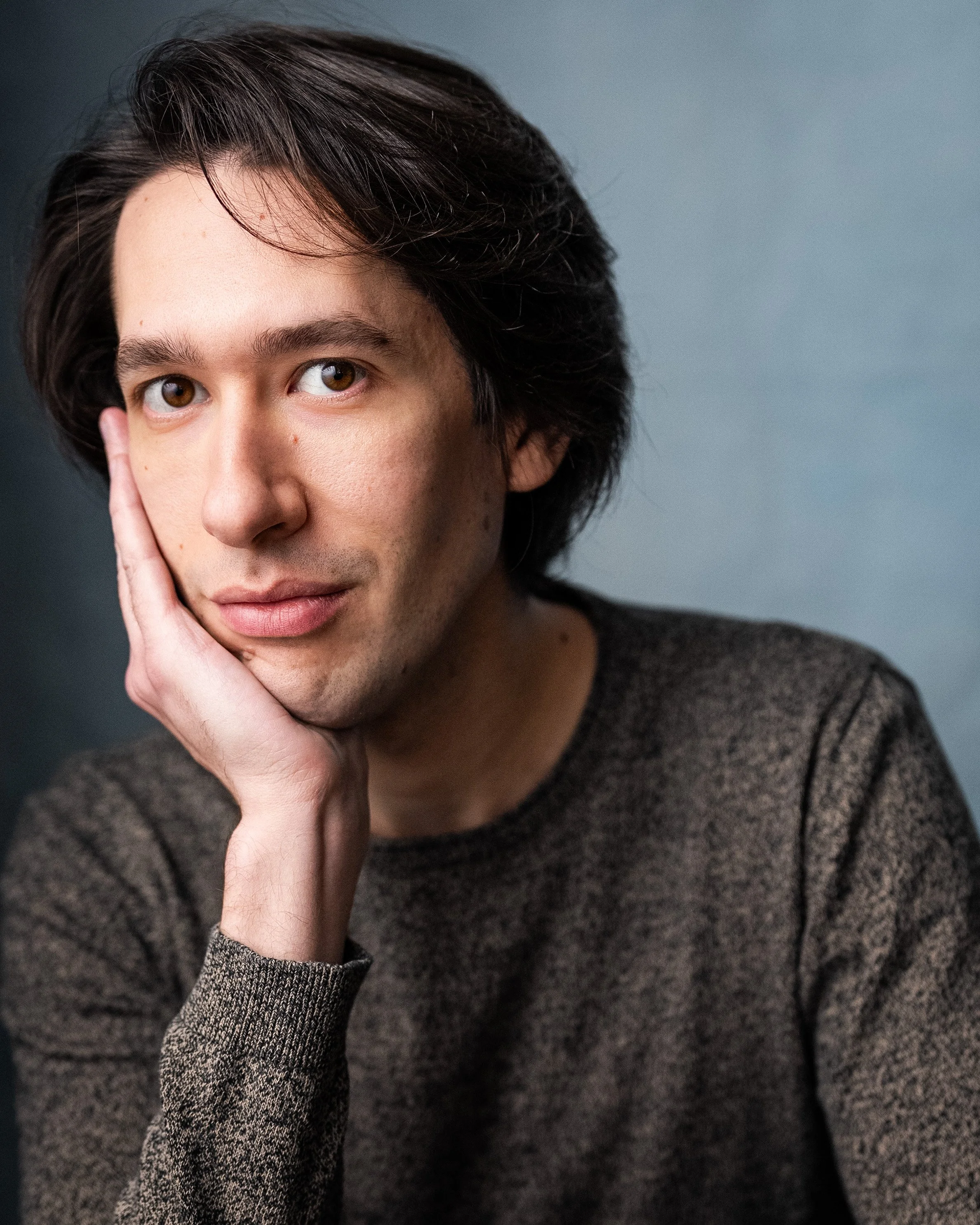 Portrait of actor Christian Pascolutti resting his head on his hand, looking directly at the camera with a slight smile, wearing a brown sweater against a plain gray background.