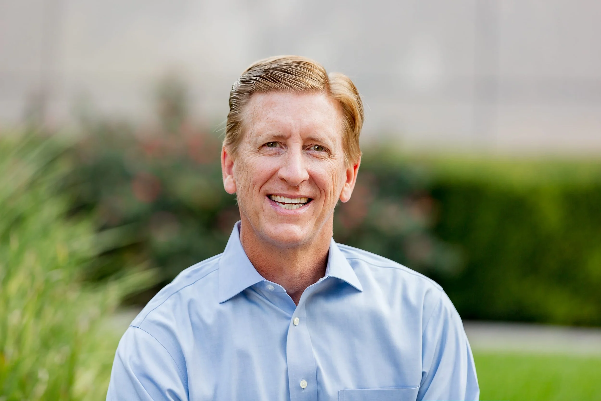 A smiling middle-aged man with light skin, blonde hair, and a blue button-up shirt standing outdoors with greenery and blurred background.