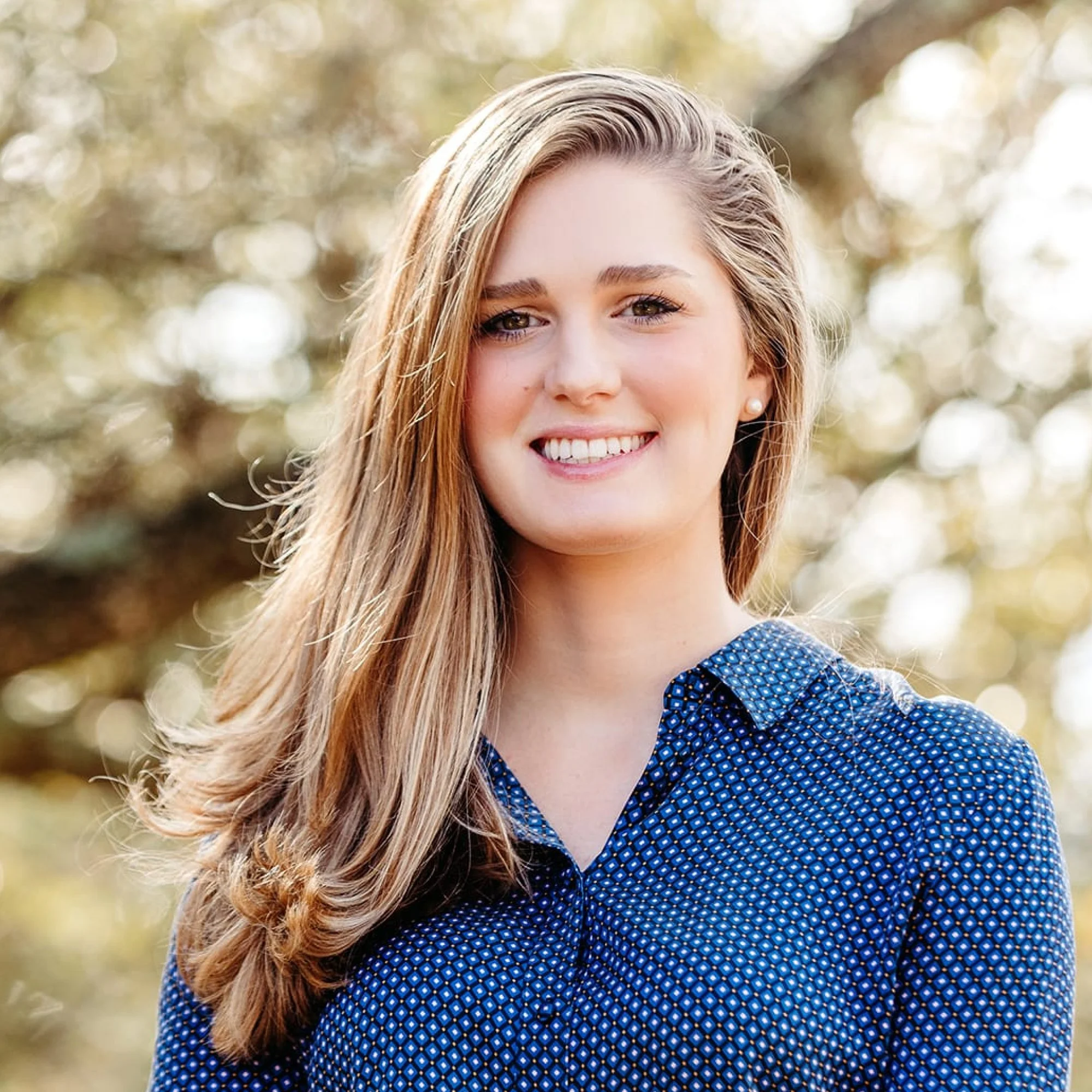 Close-up of a young woman with long blonde hair, smiling outdoors with a soft blurred background of trees and sunlight