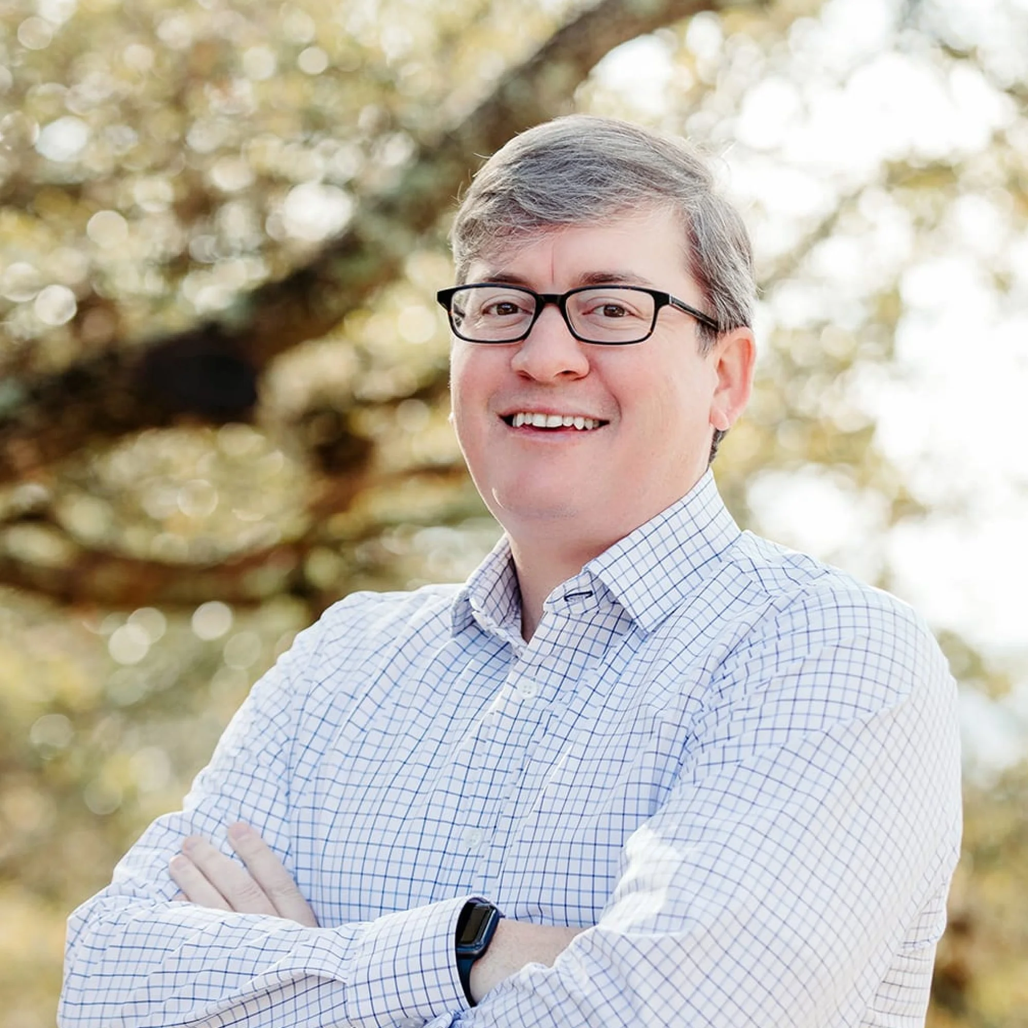 A smiling man with gray hair and glasses, wearing a checkered shirt, standing outdoors with blurry trees and sunlight in the background.