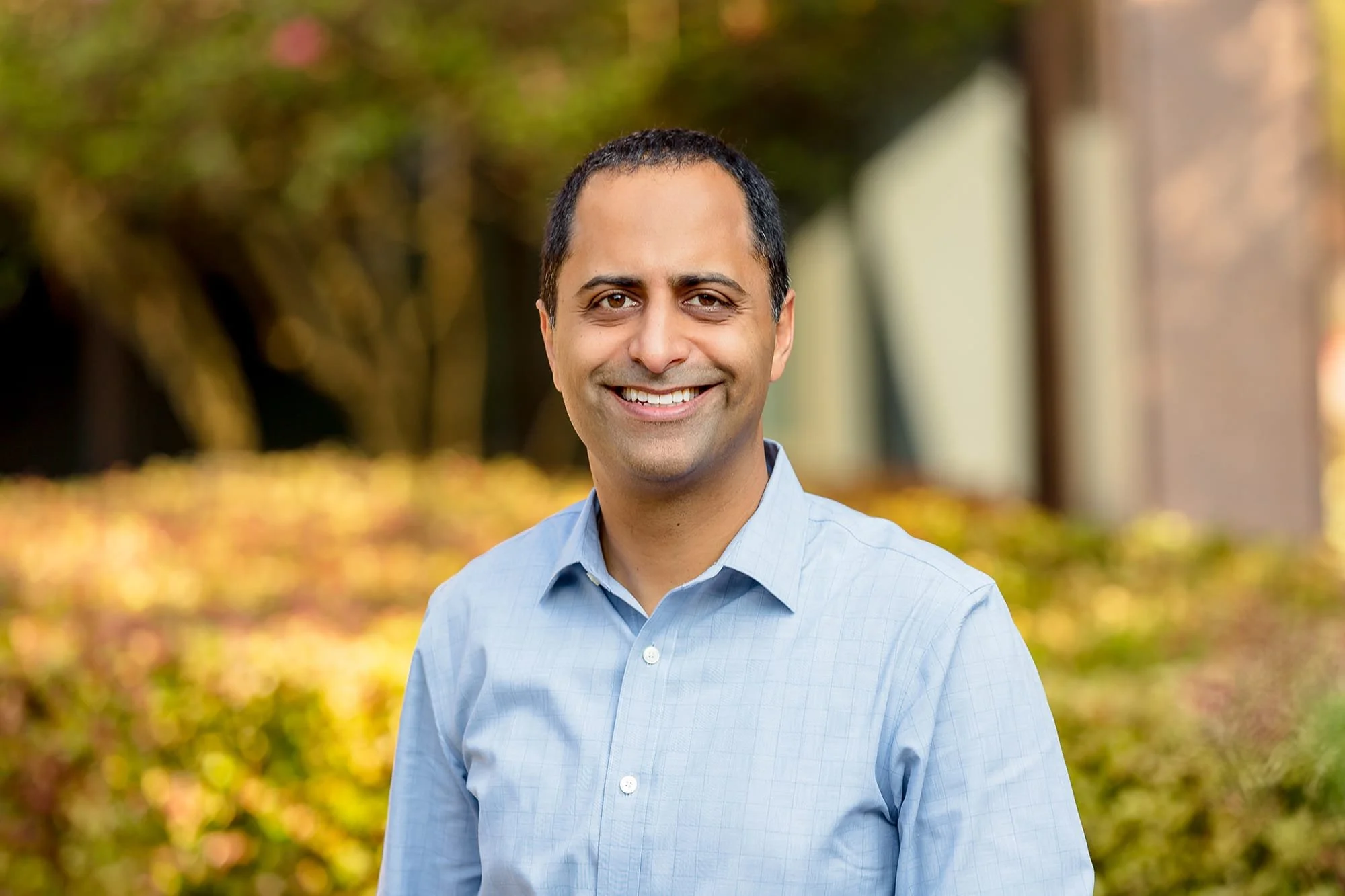 A man smiling outdoors with a blurred background of trees and buildings