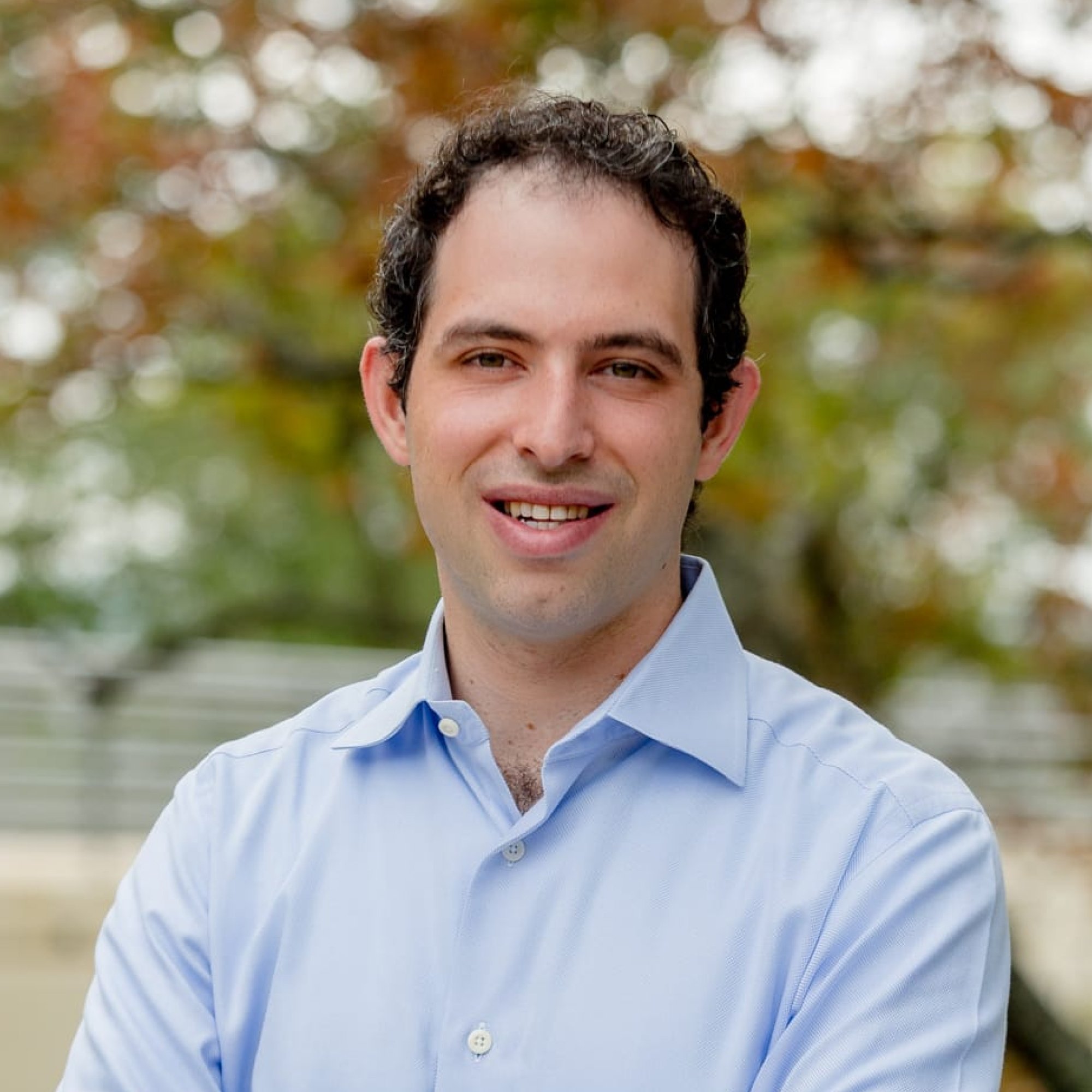 Close-up of a smiling man wearing a light blue dress shirt, outdoors with blurred fall foliage background.