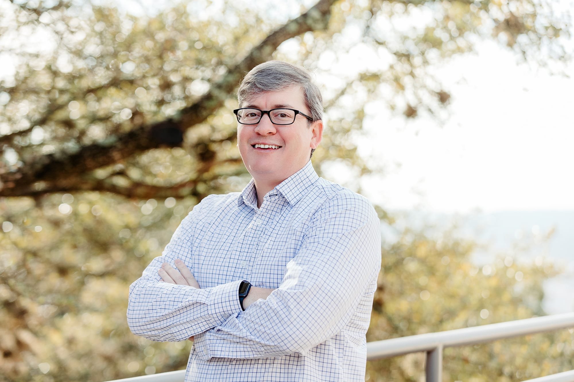 Man with glasses wearing a checkered shirt, standing outdoors with trees in the background.