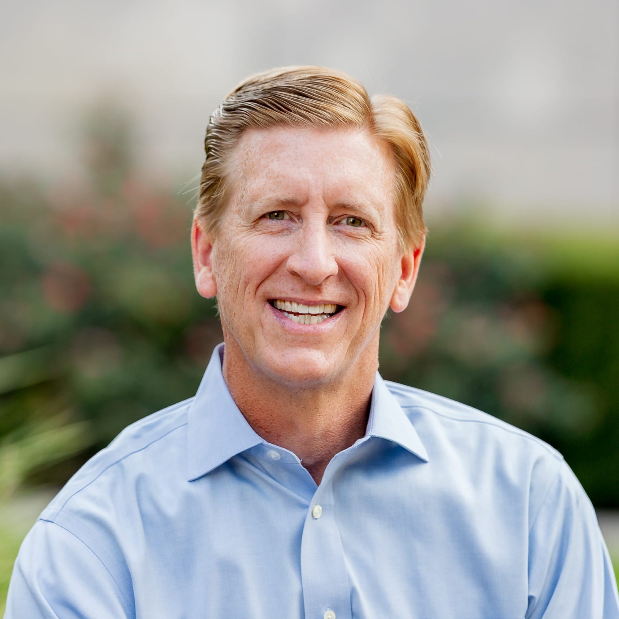 Close-up of smiling middle-aged man with red hair wearing a light blue dress shirt, outdoors with blurred greenery background.