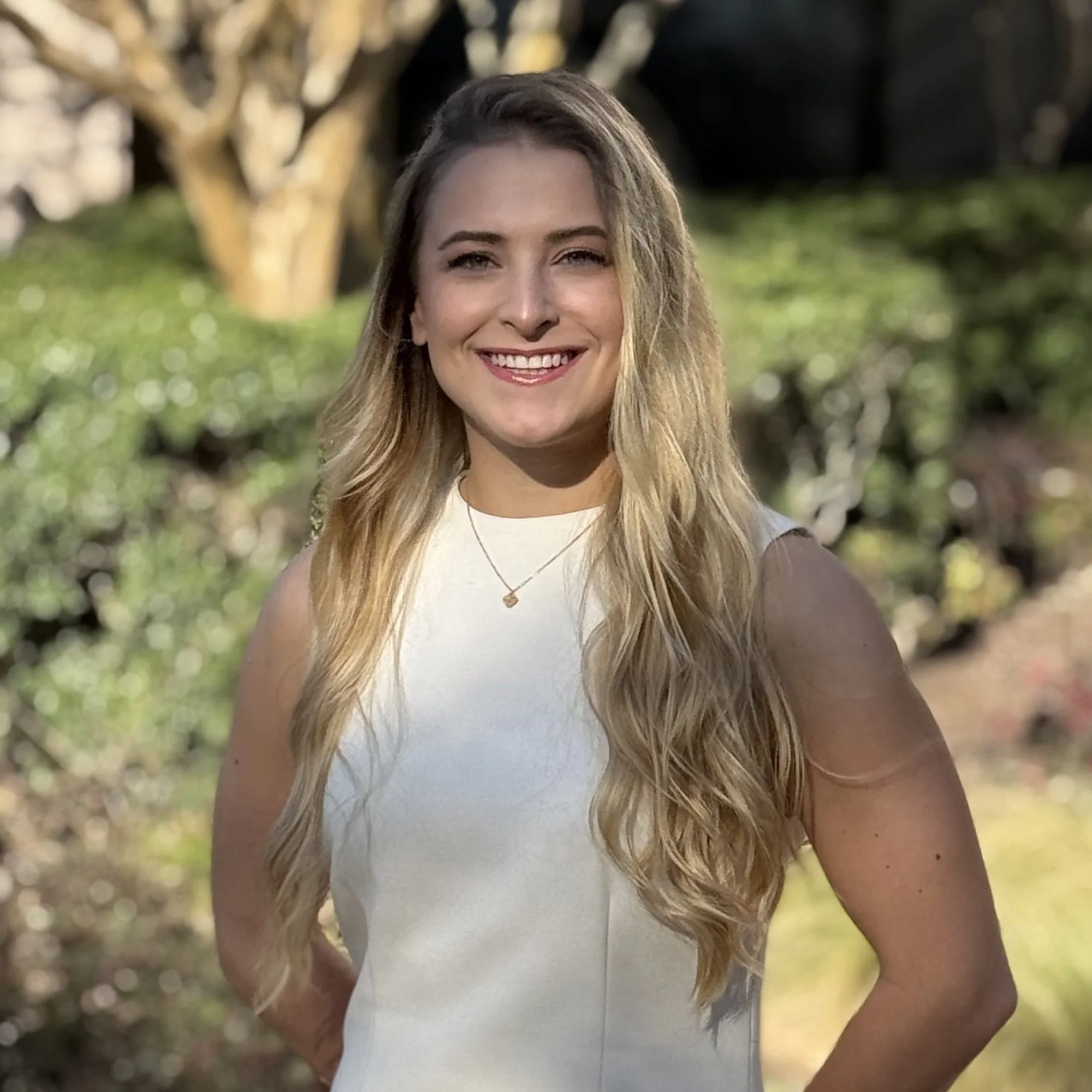 A young woman with long, wavy blonde hair, smiling, standing outdoors in sunlight with trees and greenery in the background.