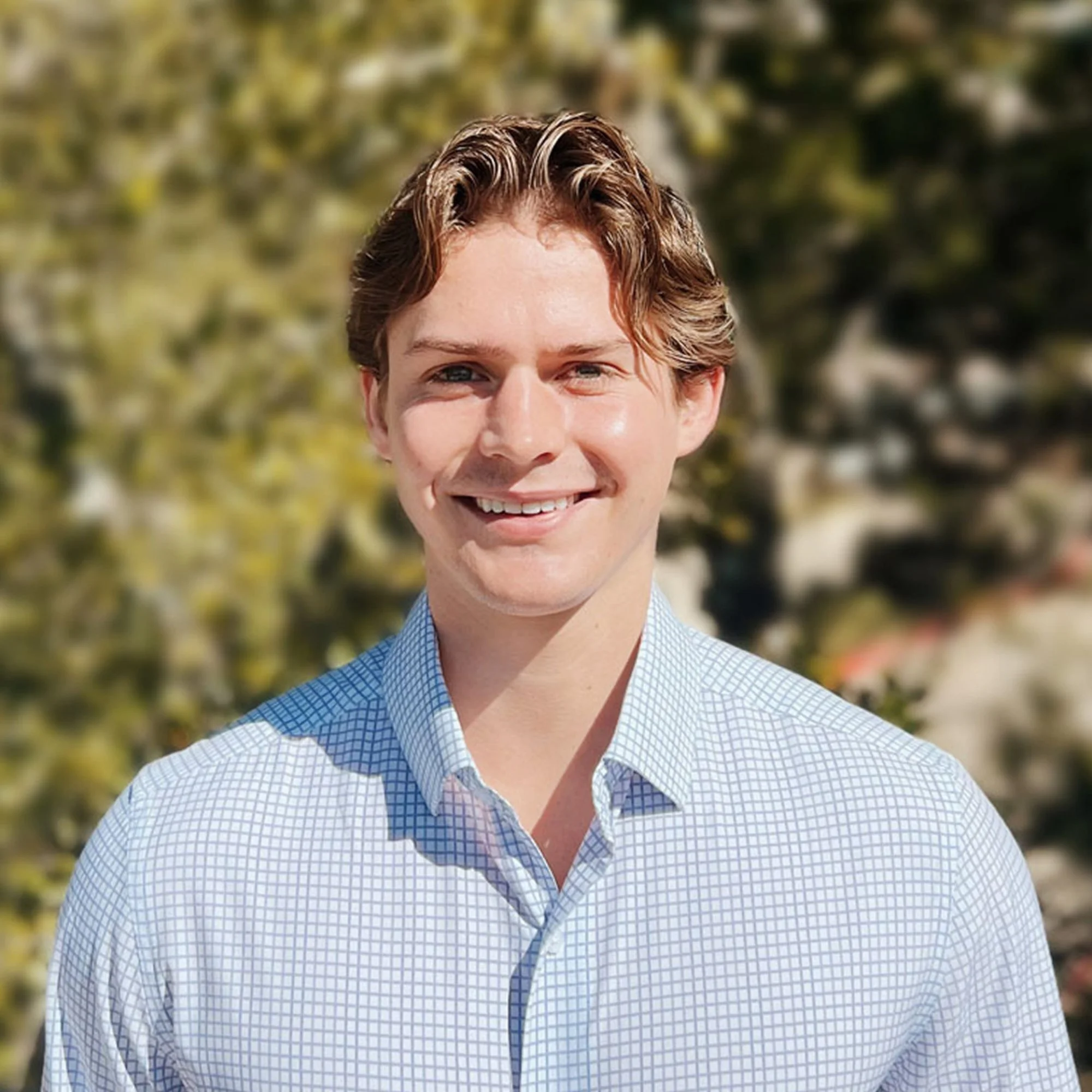 Close-up of a young man with curly hair and blue eyes, smiling, wearing a blue and white checked shirt outdoors with green foliage in the background.