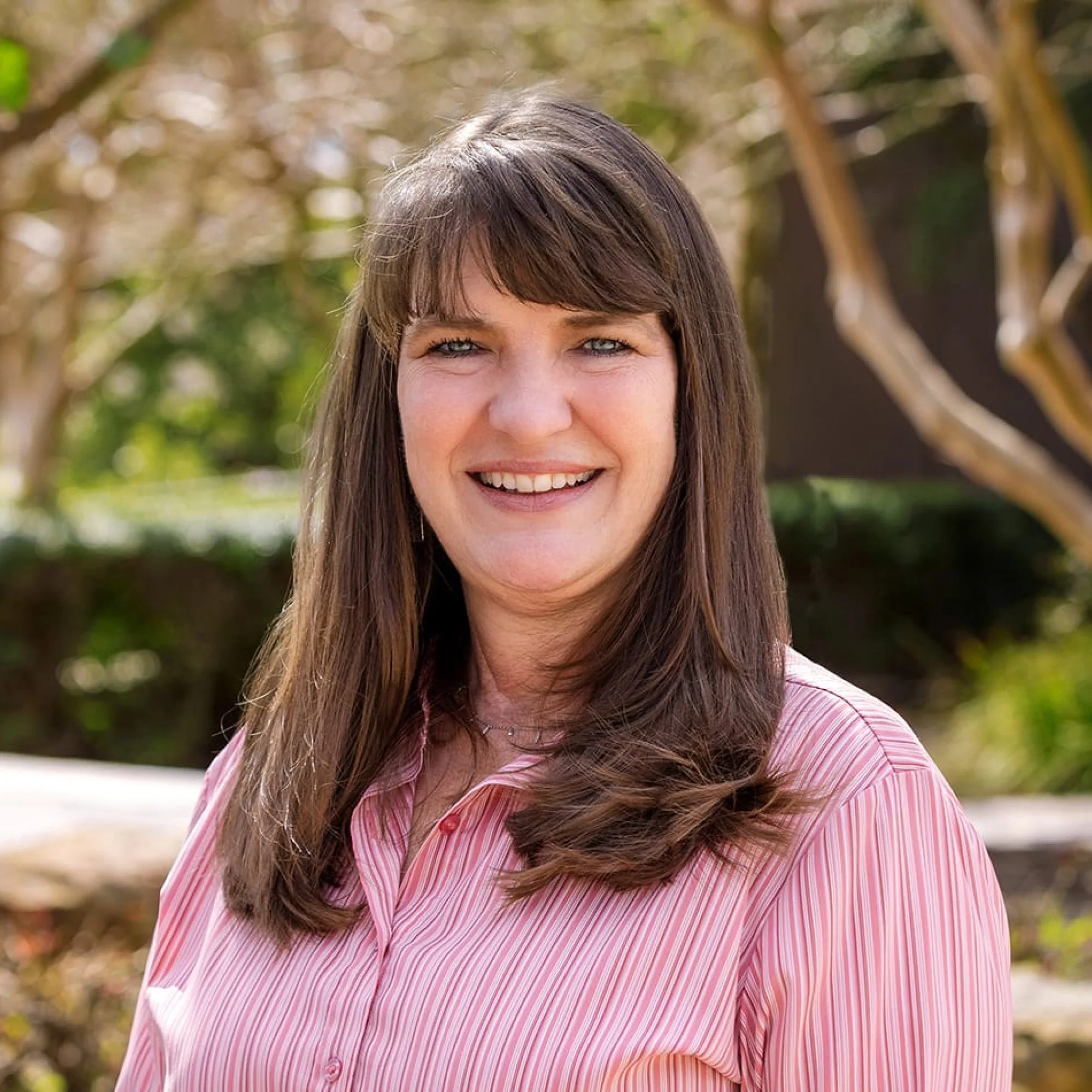 A woman with brown hair and blue eyes smiling outdoors in front of blurred trees and greenery, wearing a pink and white striped shirt.