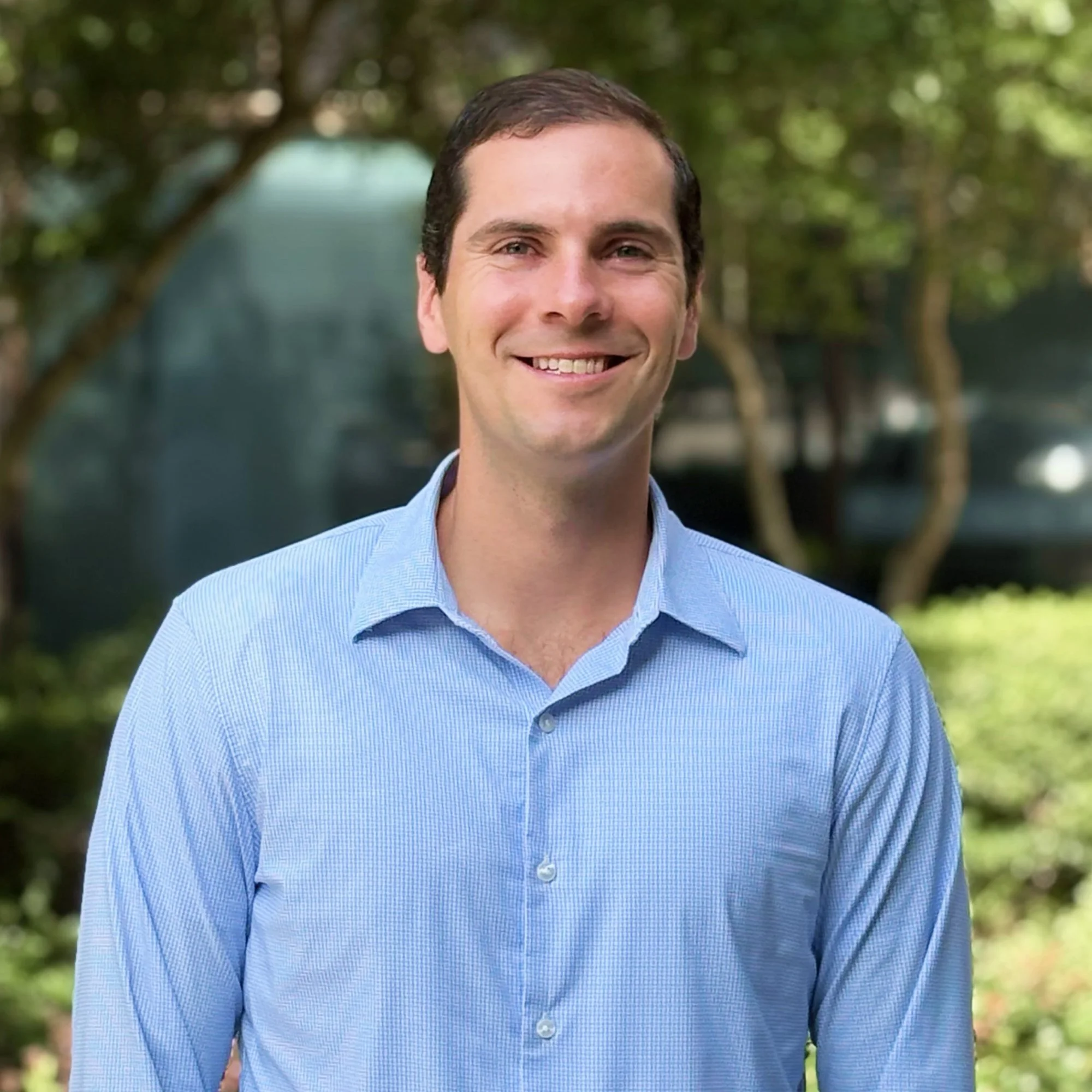A smiling man with short dark hair wearing a light blue checkered dress shirt outdoors in front of green trees.