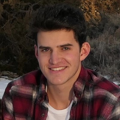 A young man with dark hair and a bright smile, wearing a red and black plaid shirt over a white T-shirt, outdoors during daytime with trees and nature in the background.