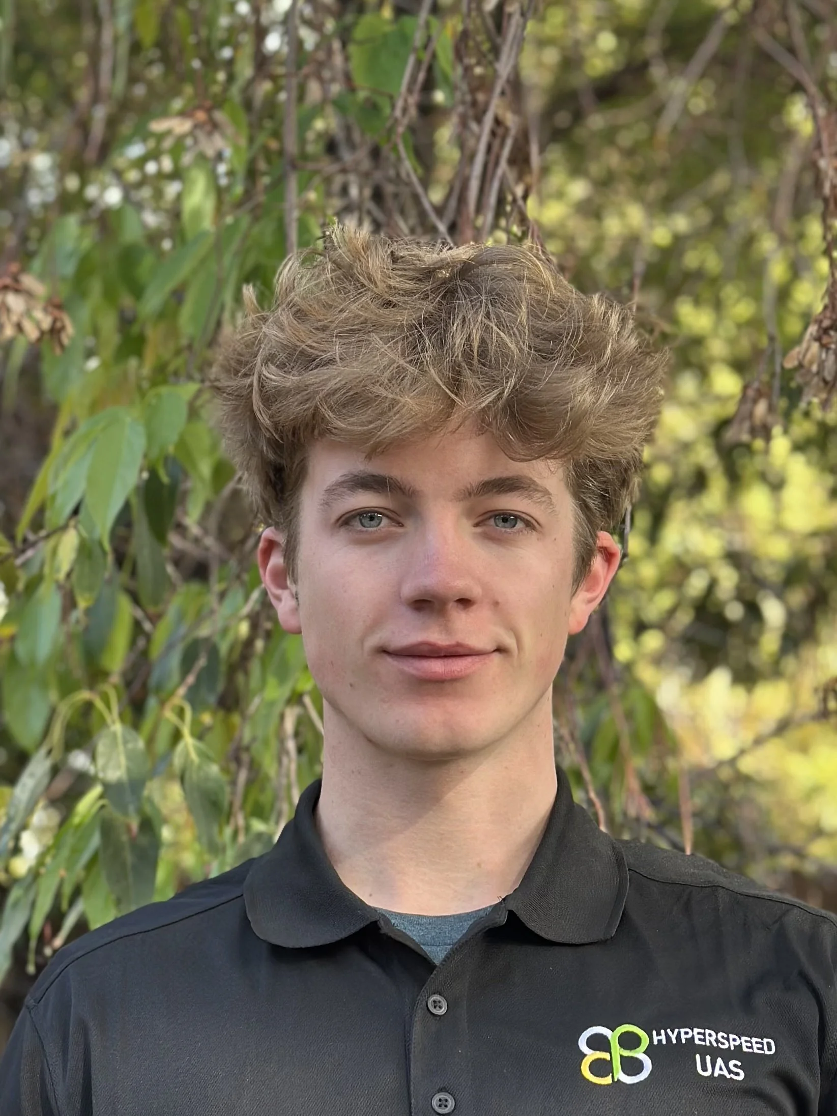 Young man with curly blonde hair and blue eyes smiling outdoors, wearing a black polo with HyperSpeed UAS logo, surrounded by green foliage.