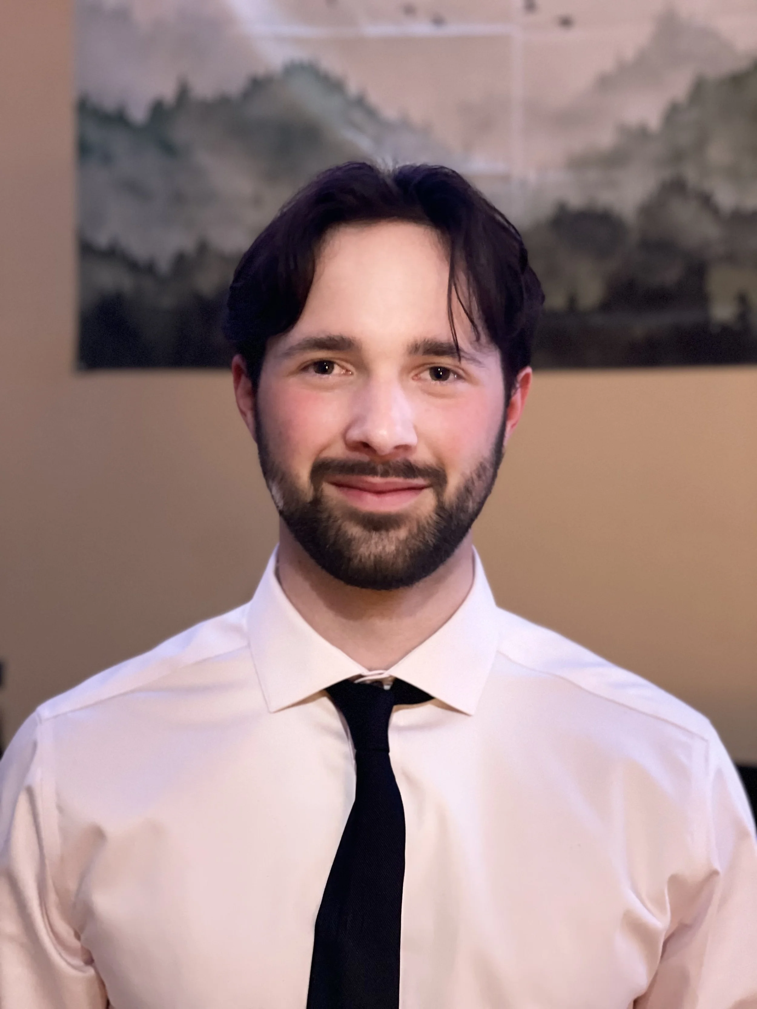 Young man with curly blonde hair and blue eyes smiling outdoors, wearing a black polo with HyperSpeed UAS logo, surrounded by green foliage.