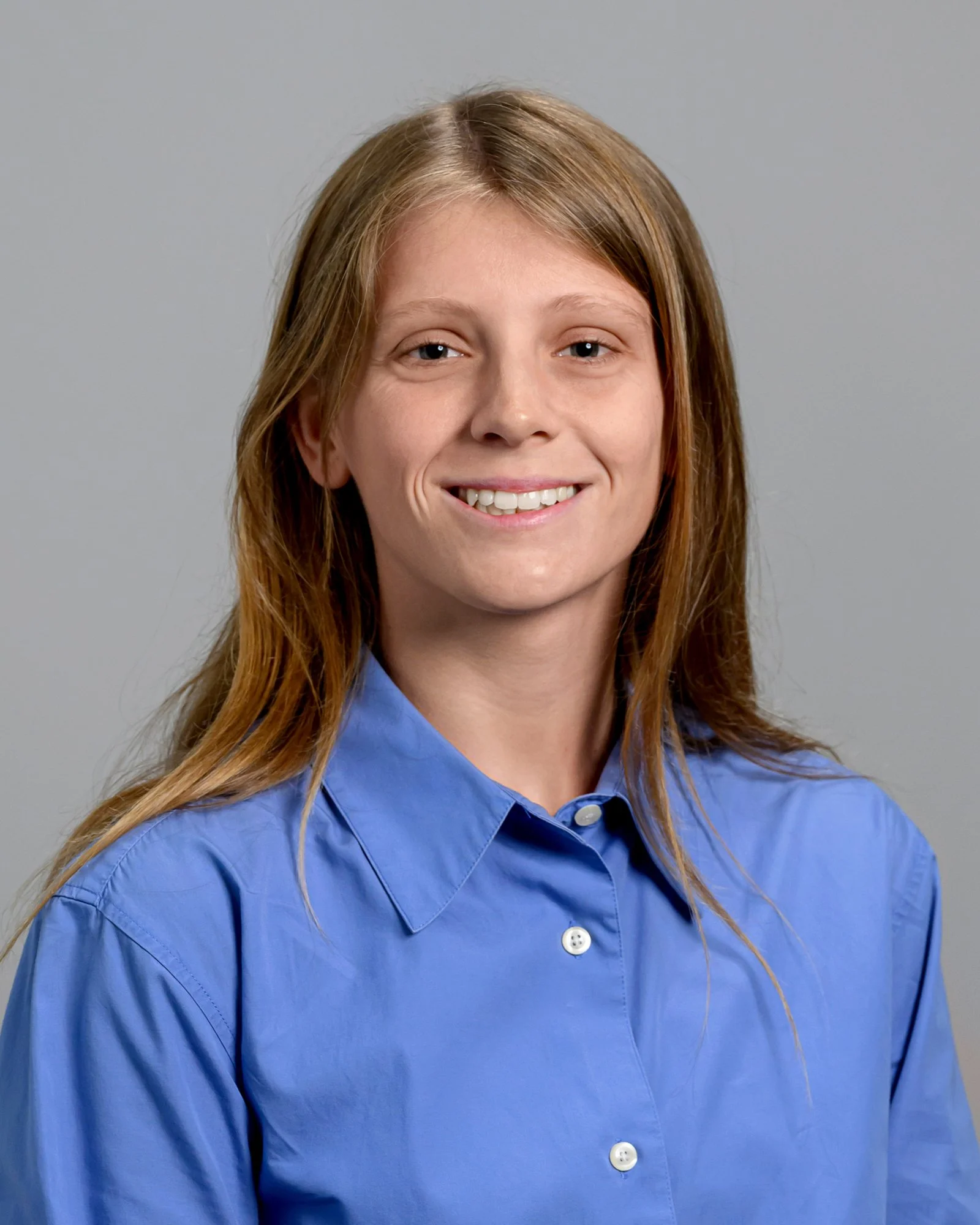 A woman with long, red hair wearing a light blue button-up shirt, smiling against a grey background.