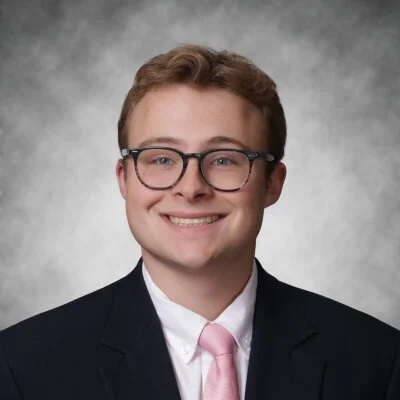 A young man in a suit with glasses, smiling for a professional portrait against a gray background.
