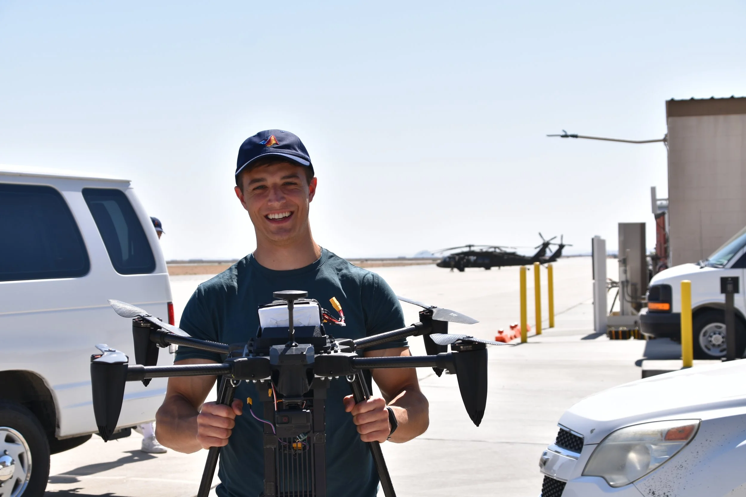 A young man with dark hair and a bright smile, wearing a red and black plaid shirt over a white T-shirt, outdoors during daytime with trees and nature in the background.