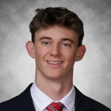 A young man with brown hair, wearing a navy suit, white shirt, and red striped tie, smiling at the camera against a gray textured background.