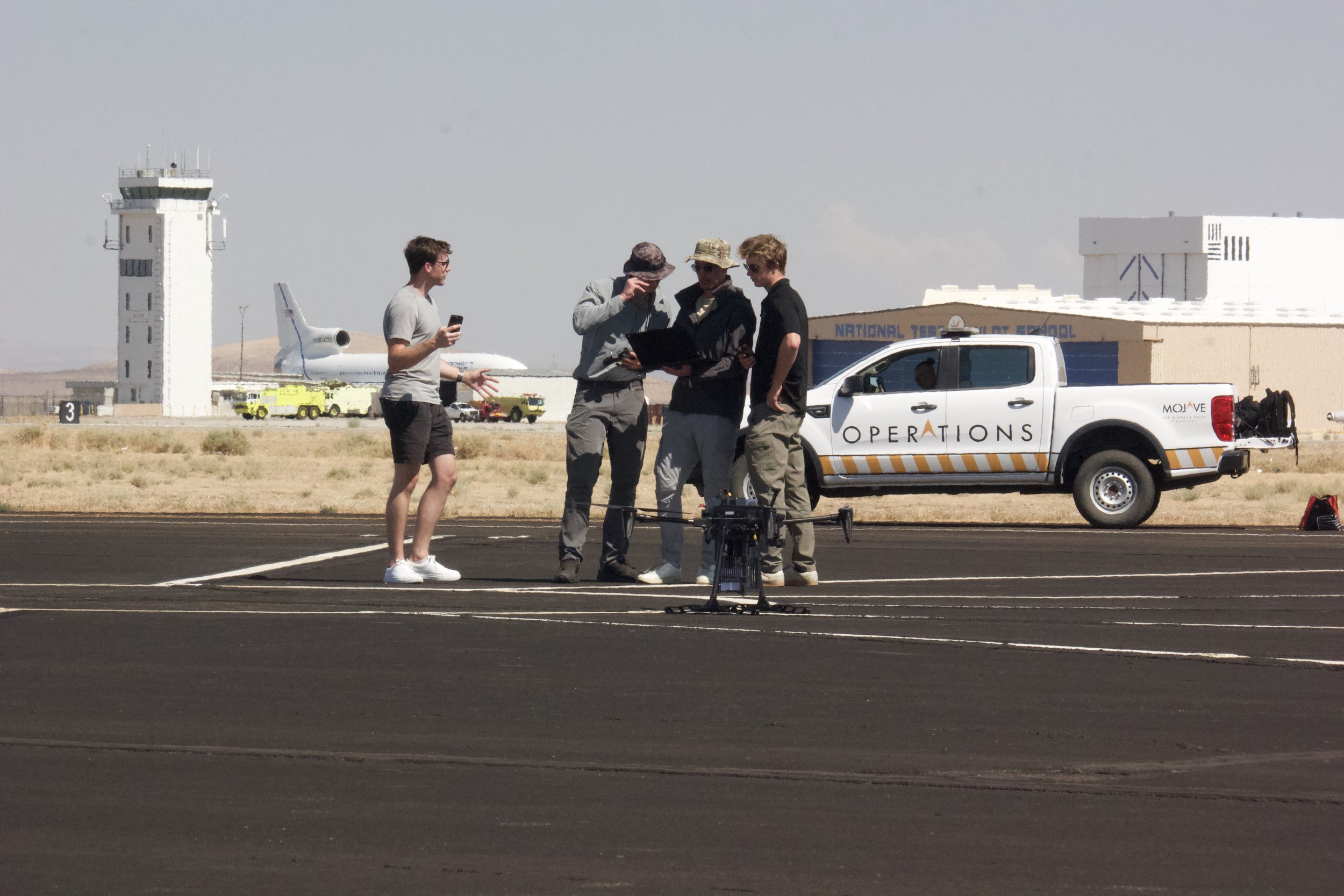 Group of four people on an airport runway, with one holding a remote control drone, discussing documents, and a laptop. A drone is on the ground near them, and an airport vehicle with 'OPERATIONS' written on the side is parked nearby. In the backgrou