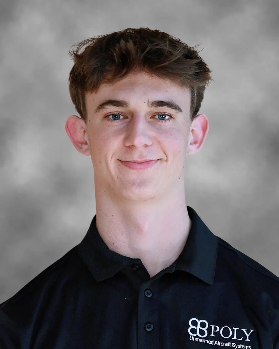 A young man with brown hair, wearing a navy suit, white shirt, and red striped tie, smiling at the camera against a gray textured background.