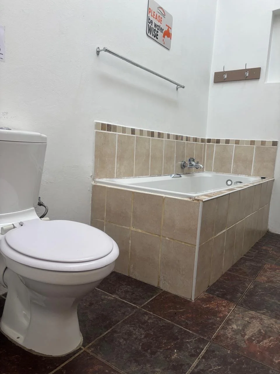 Bathroom with a toilet and a built-in bathtub, beige tile on the wall and floor, and a sign on the wall with red and black text.