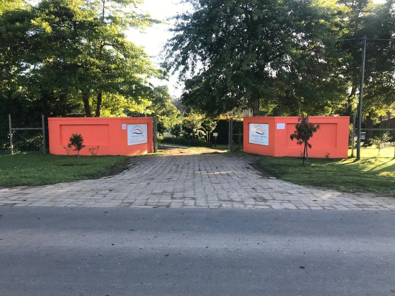 Entrance to a park with orange walls, two signs, trees, and a cobblestone pathway leading into the park.