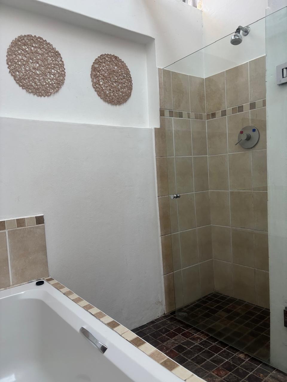 Bathroom with a partial view of a bathtub, beige tiled shower area with glass partition, decorative wall art, and a showerhead.