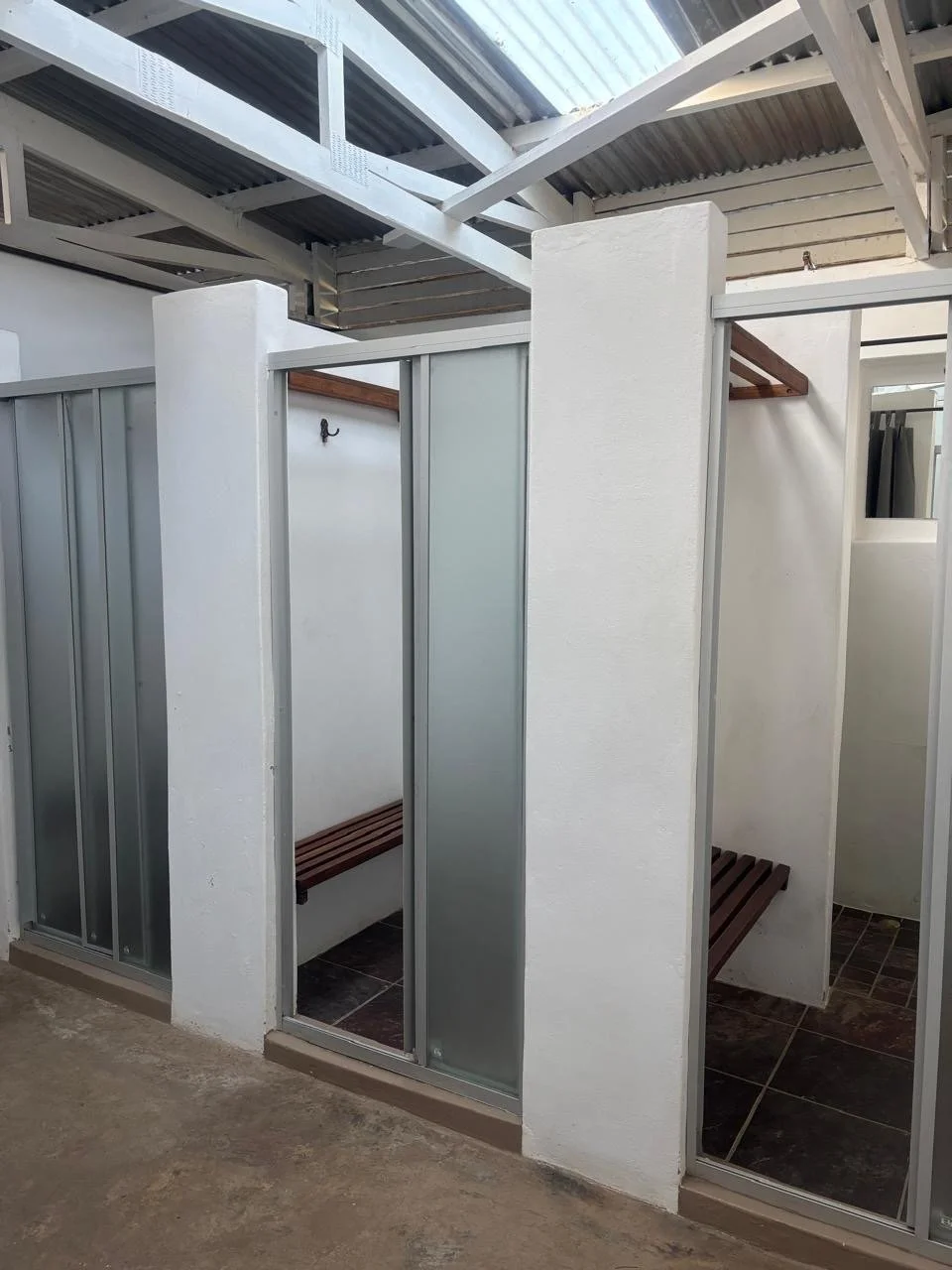 Empty outdoor shower stalls with white walls, brown wooden benches, and sliding doors, under a metal roof.
