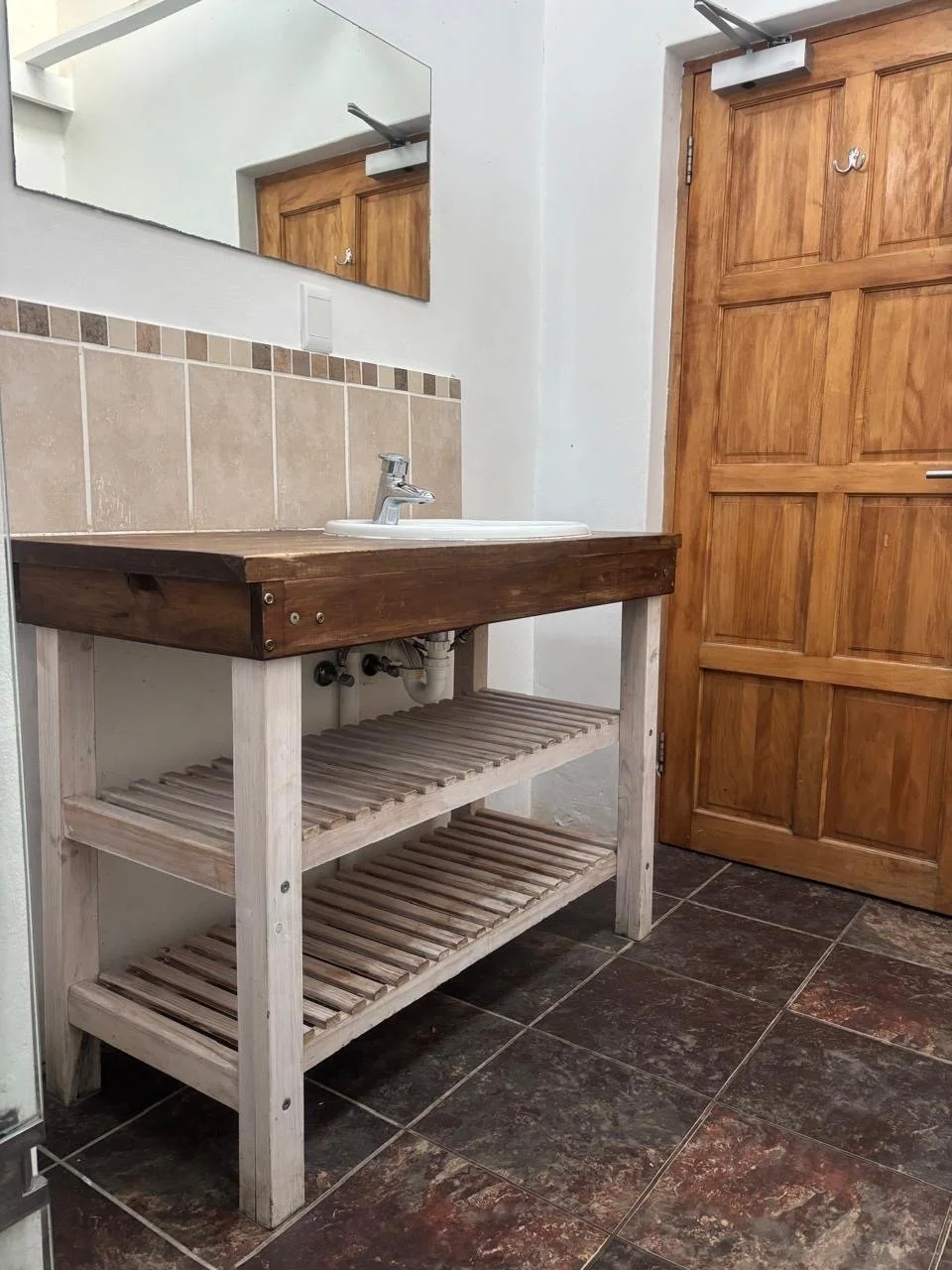 A bathroom sink with a wooden countertop and a wooden shelf underneath, a large mirror above the sink, and a wooden door in the background.