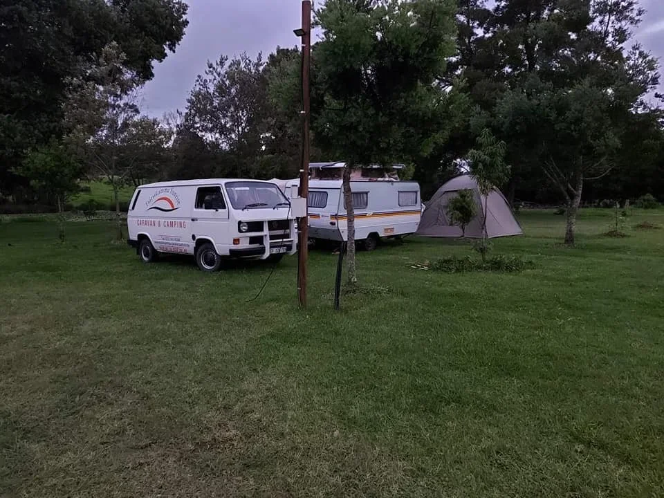 A camping scene with a white van and a travel trailer parked on a grassy field, with a tent set up nearby under trees.