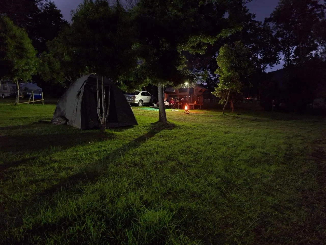 A nighttime camping scene with a tent, trees, a campfire, and parked cars in the background.