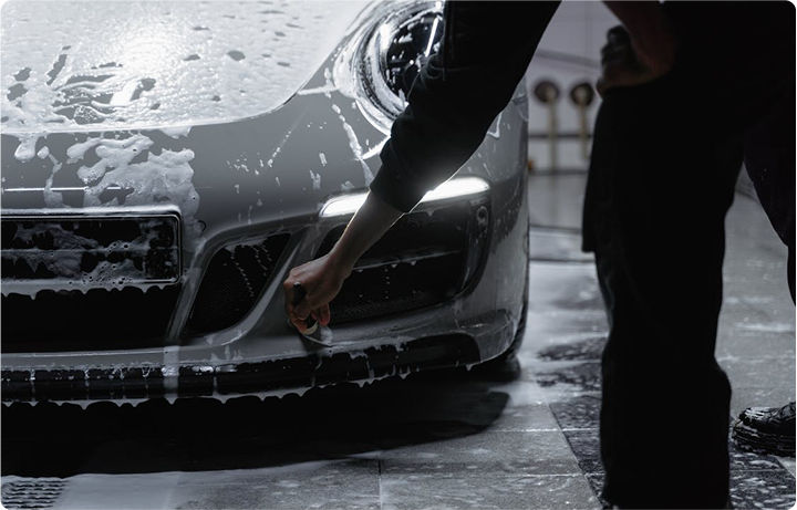 Person washing a silver sports car with soap and water in a garage, scrubbing the front bumper.